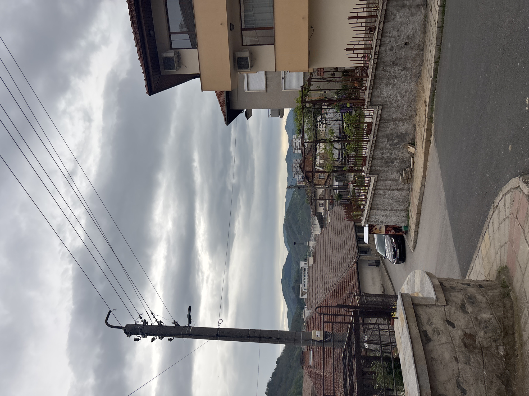 The scene depicts a street in Zlatograd, Bulgaria. The foreground contains a stone structure, partially obscured by a metal fence. Behind the fence is a house with a red tiled roof. To the left, a weathered utility pole with multiple wires extends upwards against a backdrop of heavily clouded skies. On the right side, a two-story building with beige and light grey facade features air conditioning units mounted on the walls. A stone wall runs along the side of the building, topped with a metal fence and vegetation. Beyond, the street slopes upwards. A dark-colored car with a "Coffee" sign stands on the side of the road near the top of the slope. In the distance, a mountainous landscape is visible.