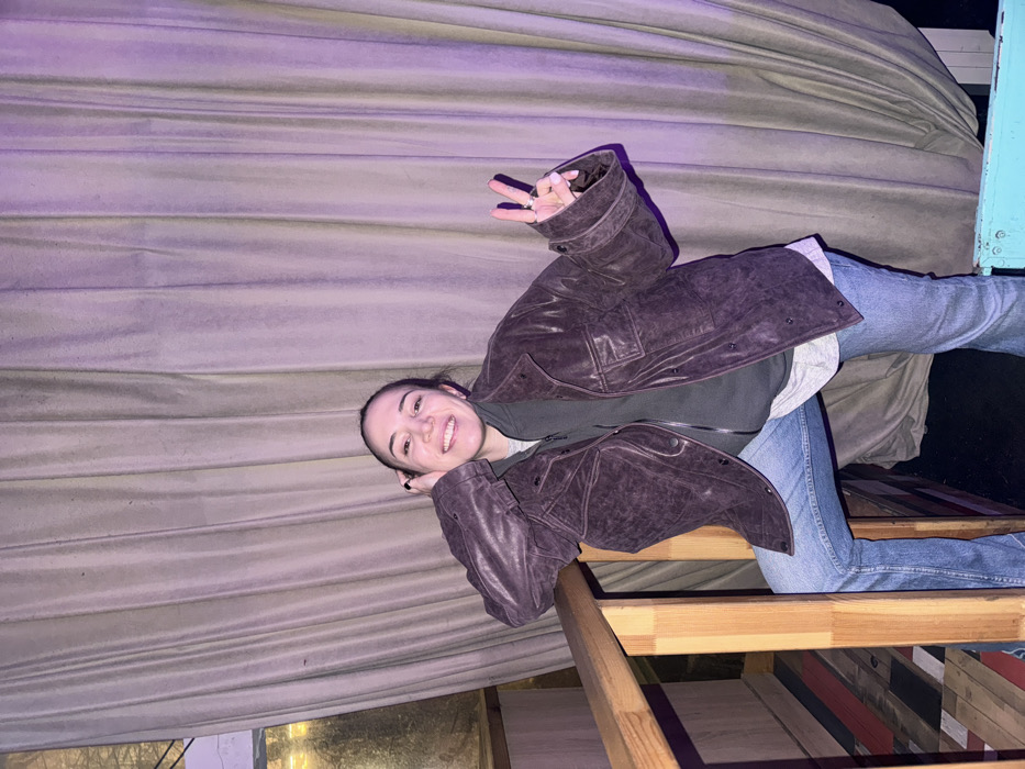 An adult female is depicted standing indoors, leaning against a light-colored wooden railing with her right forearm. She has dark hair, is smiling, and looks directly at the camera. Her right hand is positioned near her right ear, while her left hand displays a "peace" sign. She wears a dark brown leather jacket over a dark olive-green zipped hoodie, a light-colored t-shirt, and blue jeans. Her left leg appears bent, with her foot possibly resting on an elevated surface below the railing, while her right leg is extended.
The background consists largely of vertical, pleated, light-beige drapes illuminated with a purple hue on the right side. To the far left, a window or reflective surface is visible. Below the wooden railing on the left, there is a section of wall or floor with multi-colored horizontal wooden planks in shades of red, blue, and light wood. A portion of a bright blue object, possibly a chair or structural element, is visible at the bottom right. The lighting is dim and atmospheric. The environment suggests a commercial establishment such as a bar or restaurant. The specific location within the city of Moscow, Russia, is not identifiable from the visual information provided.