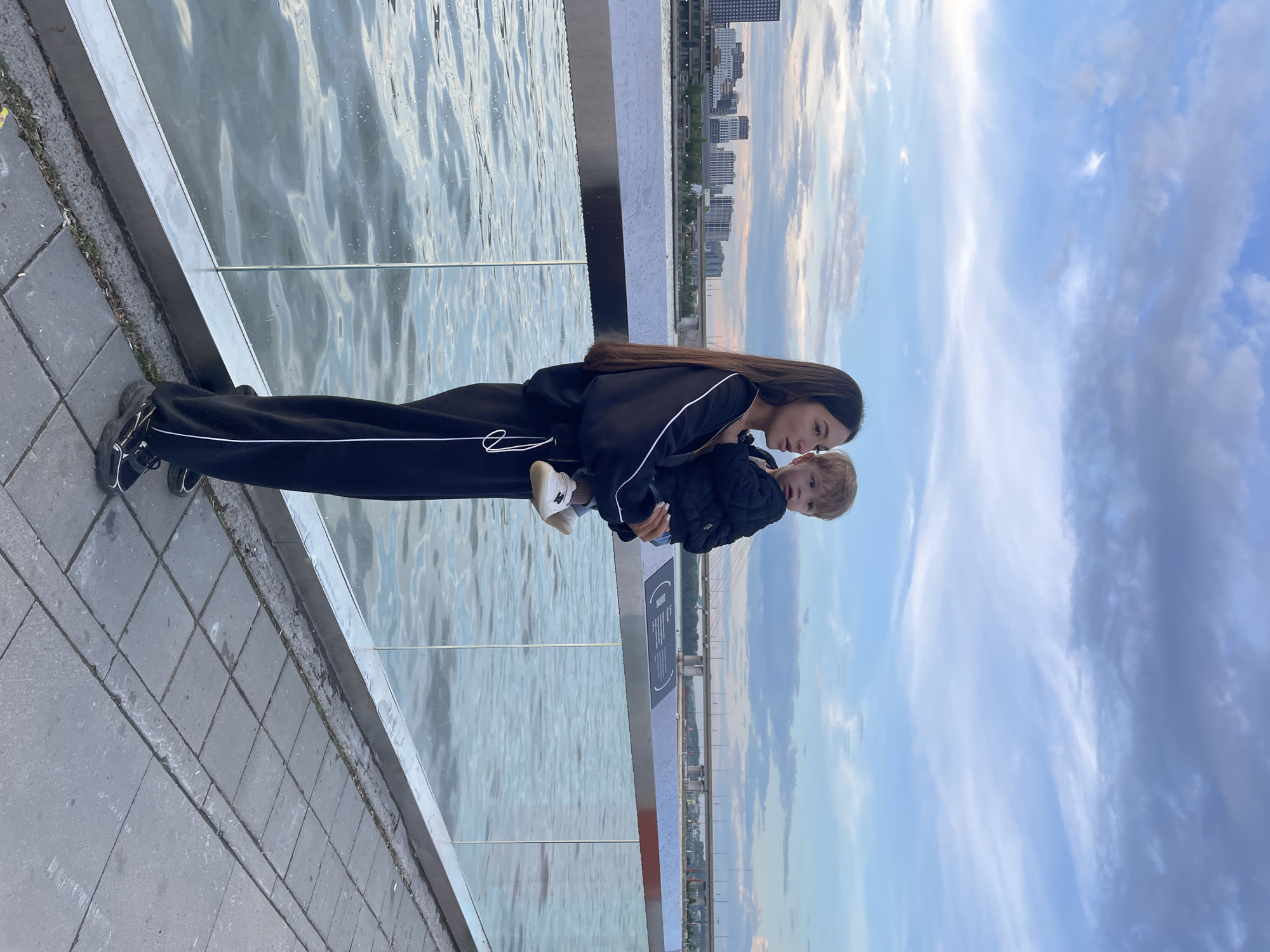 In Kazan, Russia, a young woman with long, dark hair stands on a paved walkway next to a body of water. She is wearing a black tracksuit with white piping and black sneakers. She is holding a young child, who is wearing a black jacket and white shoes. The child has blonde hair. Behind them, a city skyline is visible with buildings and a bridge. The sky is cloudy with blue patches. A glass barrier separates the walkway from the water.
