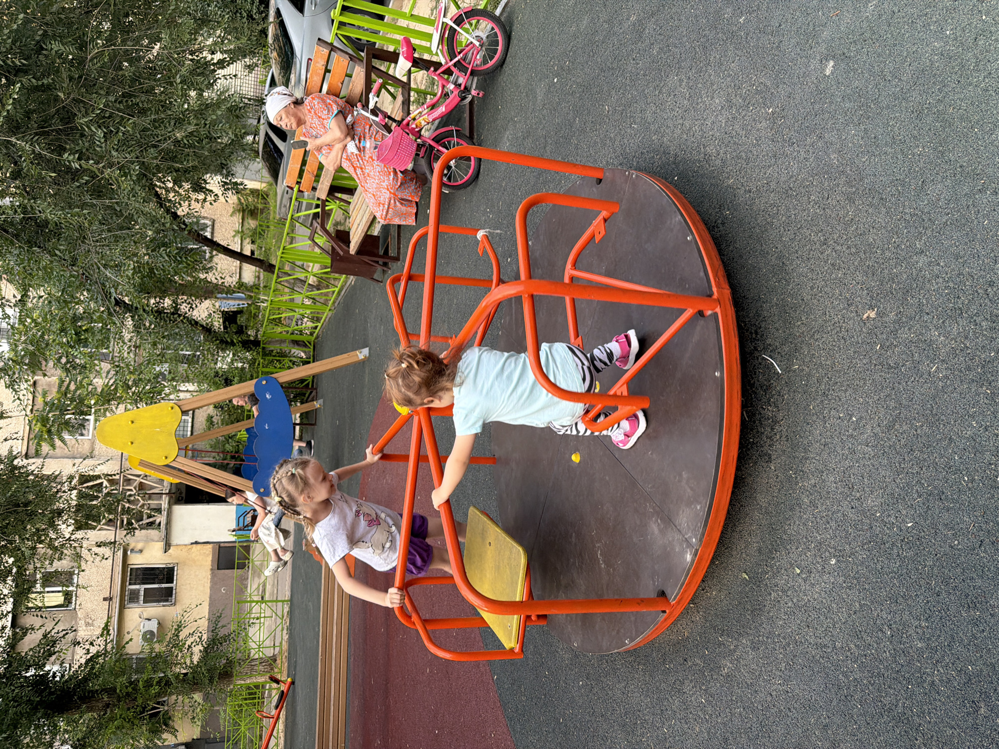 The image shows an outdoor playground in the city of Shymkent, Kazakhstan. In the foreground, two young girls are on a red and black merry-go-round. One girl, wearing a white t-shirt with a graphic and purple shorts, is standing and holding onto the railing. The other girl, wearing a light blue t-shirt and striped pants, is also on the merry-go-round, facing away from the camera. In the background, a woman wearing an orange and white patterned dress and a headscarf sits on a wooden bench, looking at her phone. A pink bicycle with a basket is parked next to the bench. Behind the bench, a light-colored car is visible. The playground area has a rubberized surface and is surrounded by a green metal fence. Trees and an apartment building are visible in the background.