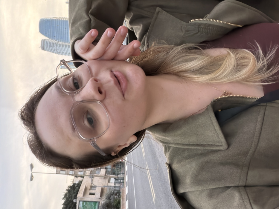 A female individual, appearing in her 20s or 30s, is captured in a self-portrait. She has light skin, brown hair with blonde highlights styled to her left side, and wears clear-framed, rectangular eyeglasses. She is dressed in an olive-green collared jacket over a reddish-brown top, with a thin gold-colored necklace visible. Her right hand, with fingers slightly splayed, rests against her right cheek. Her mouth is slightly open, and she looks directly towards the camera.
The scene is an urban outdoor environment in the city of Ramat Gan, Israel. Behind the subject, to her left, an asphalt road with white and yellow painted lines is visible, bordered by a low, dark fence. Beyond the fence, a light-colored multi-story building with a partially visible green sign is present, accompanied by trees and a tall streetlamp. In the distant background to the subject's right, two prominent, modern skyscrapers with curved, reflective facades rise against an overcast, light-colored sky. These towers are characteristic landmarks visible from various points within Ramat Gan, looking towards the Tel Aviv skyline.