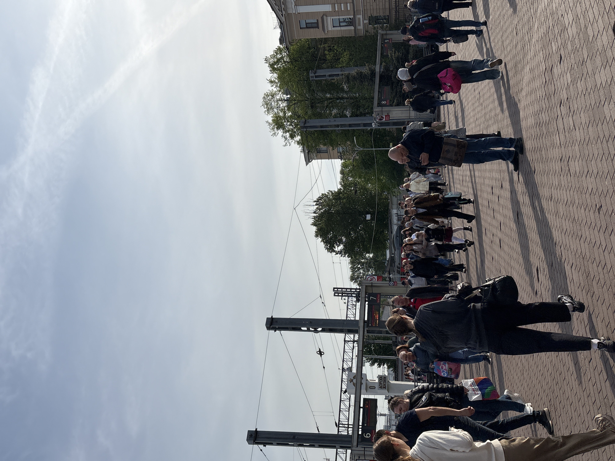 The image depicts a crowded scene in Saint Petersburg, Russia, likely near a train station or public transport hub. Numerous people are walking or standing on a brick-paved area. Overhead electrical lines and metal support structures are visible. Signs with numbers (6, 7, 8) are present, potentially indicating platform numbers. A monument or memorial is partially visible in the background, featuring a red star. The sky is overcast. The image shows people moving in various directions, and the overall impression is of a bustling public space.