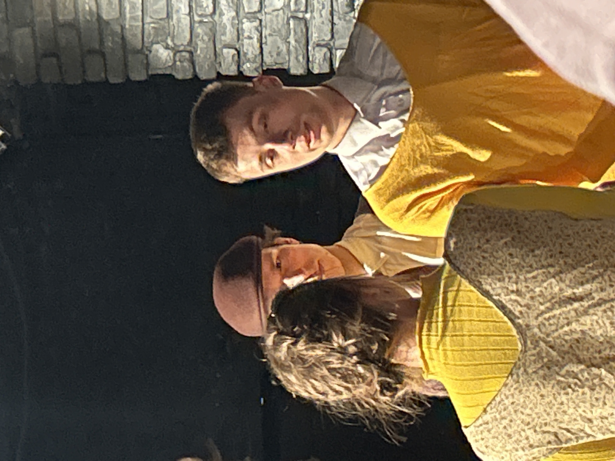 Three people are visible in the image. The person at the top wears a yellow tunic over a white collared shirt. The person in the middle wears a brown cap and light-colored shirt. The person at the bottom has brown hair and wears a yellow shirt with a floral pattern. The environment is dark, with a brick wall visible in the upper left corner. The location is Unknown, Unknown.