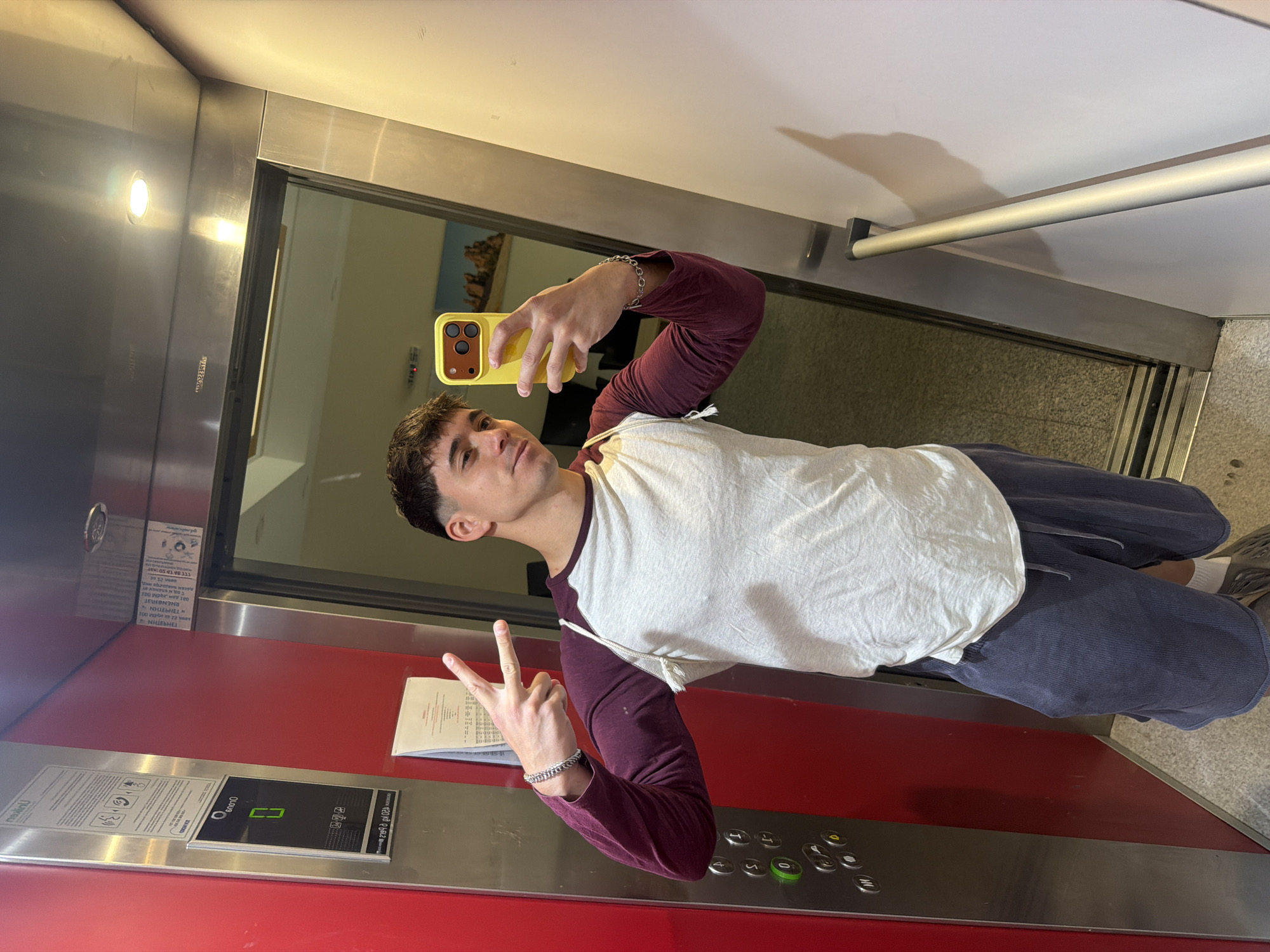 A young man poses for a selfie inside an elevator. The elevator's interior has a red and silver panel with a control panel featuring buttons and a digital display. A piece of paper is attached to the panel. The man is holding a yellow phone and making a peace sign with his fingers. He is wearing a white t-shirt with burgundy sleeves, dark pants, and a silver bracelet. The elevator doors are partially open, revealing a hallway with a painting on the wall. The exact location in Sofia, Bulgaria, cannot be determined from the image.