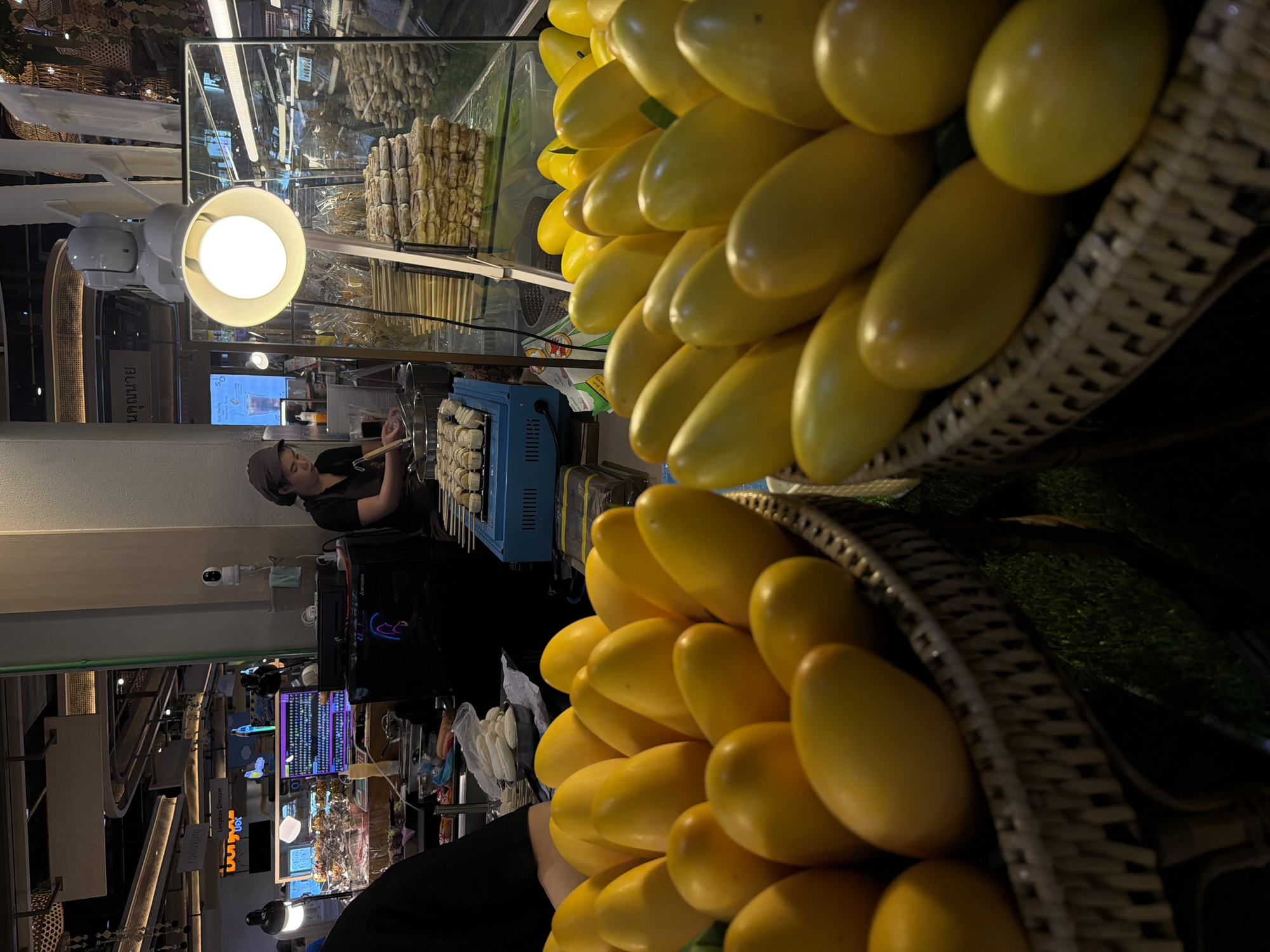 The image depicts a food stall in Chiang Mai City Municipality, Thailand. The stall has two large baskets filled with yellow, oval-shaped objects, possibly fruits or decorative items, dominating the foreground. In the background, a woman is grilling food on skewers on a small blue gas stove. She is wearing a dark shirt and a head covering. Above her, there is a lit lamp and a glass display case filled with food items. The stall is positioned in a bustling indoor market or food court setting, with glimpses of other stalls, signage, and general activity visible in the background. A security camera is mounted to a wall.