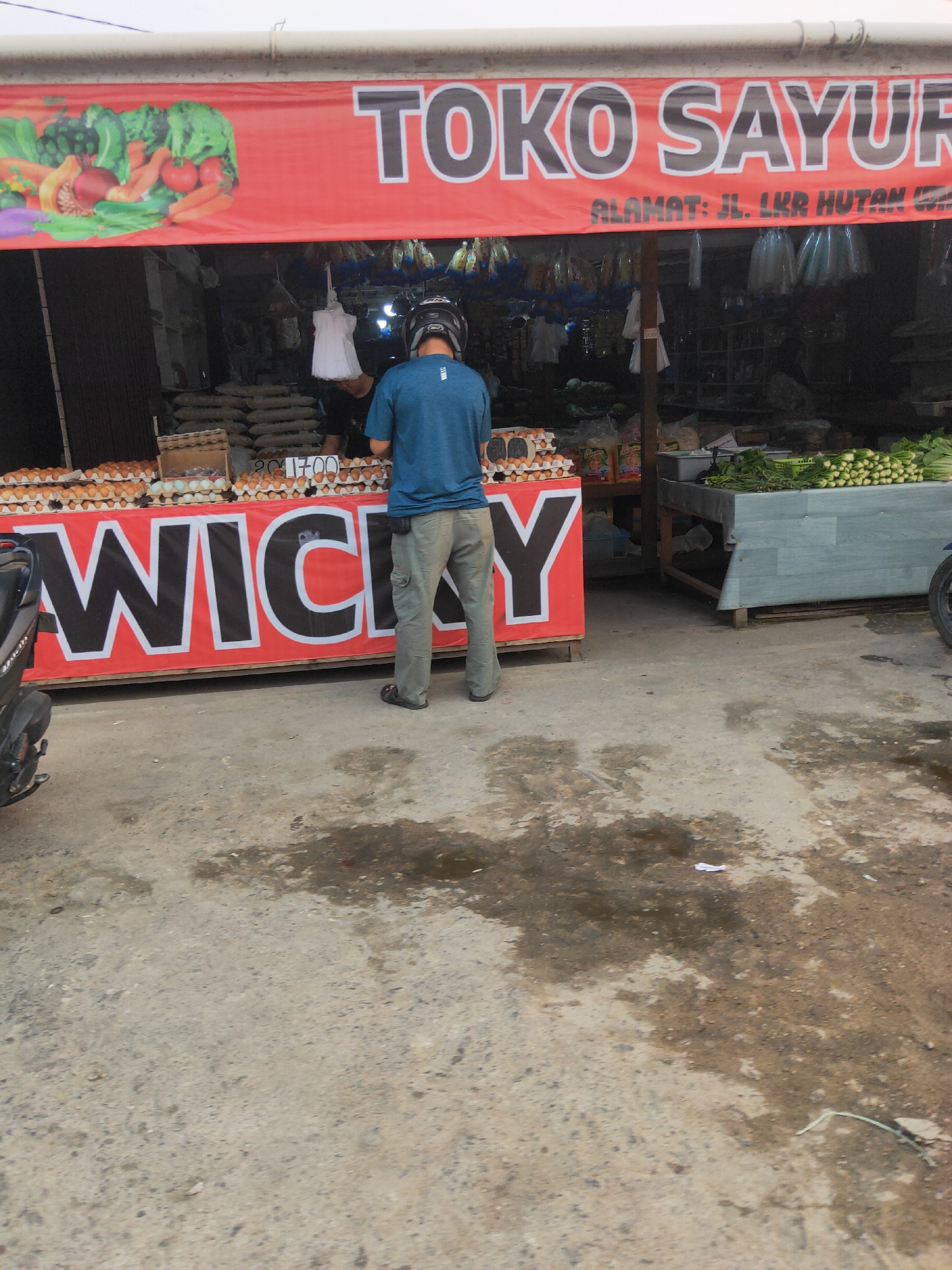 A full shot shows a man wearing a blue t-shirt and gray pants standing in front of a fruit and vegetable stand in Sintang, Indonesia. The stand is marked with a large red banner displaying the words "TOKO SAYUR" and "ALAMAT: JL. LKR HUTAN (W..". The man, facing away from the camera, is wearing a helmet and appears to be interacting with a vendor, though the vendor is not fully visible. To the left of the man, there are stacks of eggs on display, and to his right, there are various vegetables. The ground in front of the stand is paved and shows signs of being wet. The overall impression is a busy market scene.