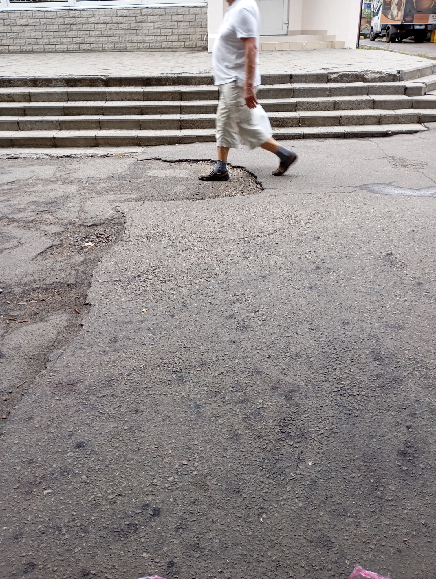A person is walking across a cracked asphalt surface in Horishni Plavni, Ukraine. The person is wearing a white polo shirt, beige shorts, dark socks, and brown shoes. They are walking away from a set of concrete stairs and towards the foreground of the image. The stairs appear weathered and are bordered by a tiled walkway. In the background, a building with a white facade and a truck are visible. The asphalt in the foreground is broken and uneven, with visible debris.