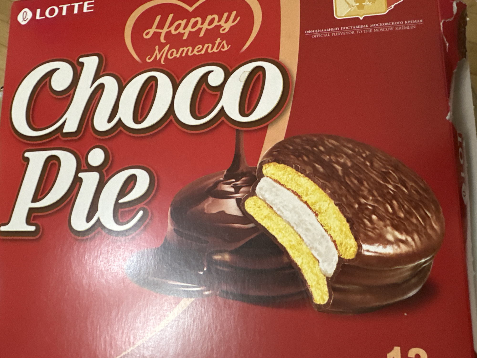 A close-up, eye-level, full-frame shot features a red box of Lotte Choco Pie. The box displays prominent white text with brown outlines, spelling out "Choco Pie." To the right of the text, a graphic depicts a partially unwrapped Choco Pie with layers of yellow cake, white cream, and dark chocolate coating. Below the "Choco Pie" text, "Happy Moments" is written in a cursive font within a circular design. Along the right edge of the box, "LOTTE" is printed vertically in white text. At the bottom right, a small portion of text in Russian and English reads "OFFICIAL PURVEYOR TO THE MOSCOW KREMLIN." The background is a plain red surface, likely the box itself. No people or specific Yerevan locations are visible.
