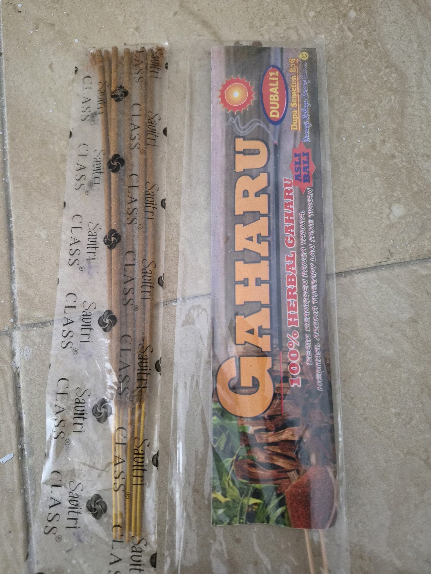 The image shows two packages of incense sticks on a tiled surface. The package on the left contains several thin, brown incense sticks bundled together and sealed in a clear plastic wrapper with the repeating text "Savitri CLASS". The package on the right is a taller, wider wrapper with a colorful label featuring the word "CAHARU" prominently in large orange letters. Below "CAHARU", smaller text reads "100% HERBAL GAHARU" and then "MEDITASI AROMA THERAPY LAN SANE TIOSAN". To the upper right of "CAHARU", there are smaller text elements including "DUBALI1", "Dupa Semeton Boli", and "Dharma Rahaya Sarang". There is also a small circular emblem with a red sun-like graphic and a small smiley face. The incense sticks themselves are visible through the clear plastic of both packages. The tiled surface appears to be an outdoor or semi-outdoor area, with some small white specks scattered across it. There are no people or discernible actions depicted in the image. The location is specified as Srimahi, Indonesia, but this information is not visually evident from the image itself.