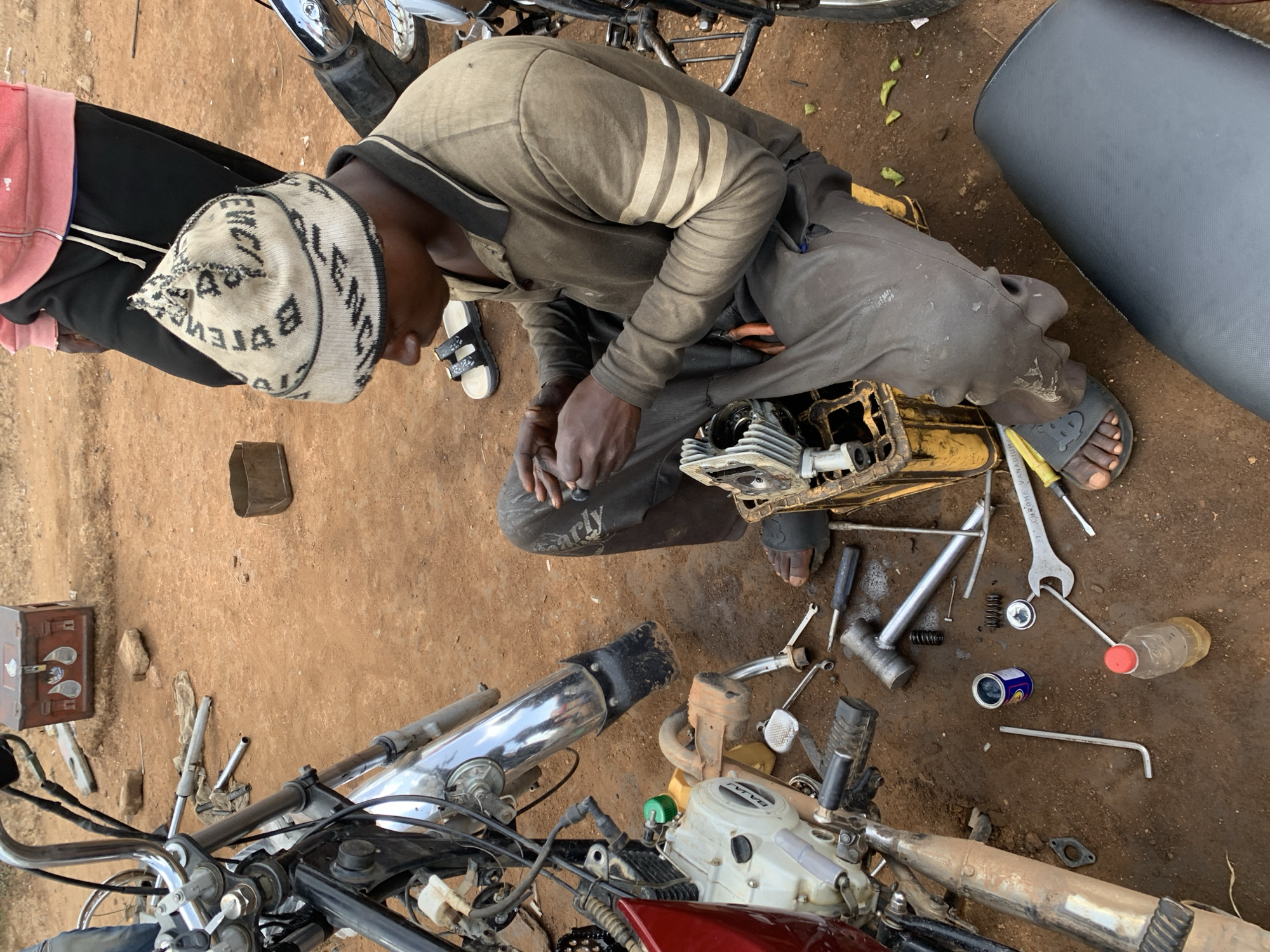 This media file shows a top-down view of a person working on a motorcycle engine in an outdoor setting. The ground is made of reddish-brown dirt, characteristic of a dry, possibly rural area. Scattered tools and motorcycle parts are visible on the ground, including wrenches, screwdrivers, springs, and what appears to be a disassembled engine block. A partially visible motorcycle seat, a pink hooded jacket, and black pants suggest the person is wearing casual work attire. The person, a man, is wearing a patterned beanie and is engaged in mechanical work, holding tools with his hands.
In the background, parts of two motorcycles are visible. One, closer to the foreground, has a chrome exhaust pipe. The other, partially obscured by the seated person, shows a wheel and frame. There's a small, dark rectangular object on the ground, possibly a metal box. A plastic bottle with a red cap and a blue and silver can are also present amongst the tools. The overall scene appears to be a workshop or a roadside repair area. The lighting suggests it's daytime, possibly late morning or afternoon. There are no clear indicators of specific weather conditions beyond it being an outdoor environment.
The visible text includes "BALENCIAGA" on the beanie the person is wearing. The engine block has some labeling, but it is not clearly legible. The overall impression is one of diligent work and self-sufficiency, with the individual focused on repairing the vehicle.