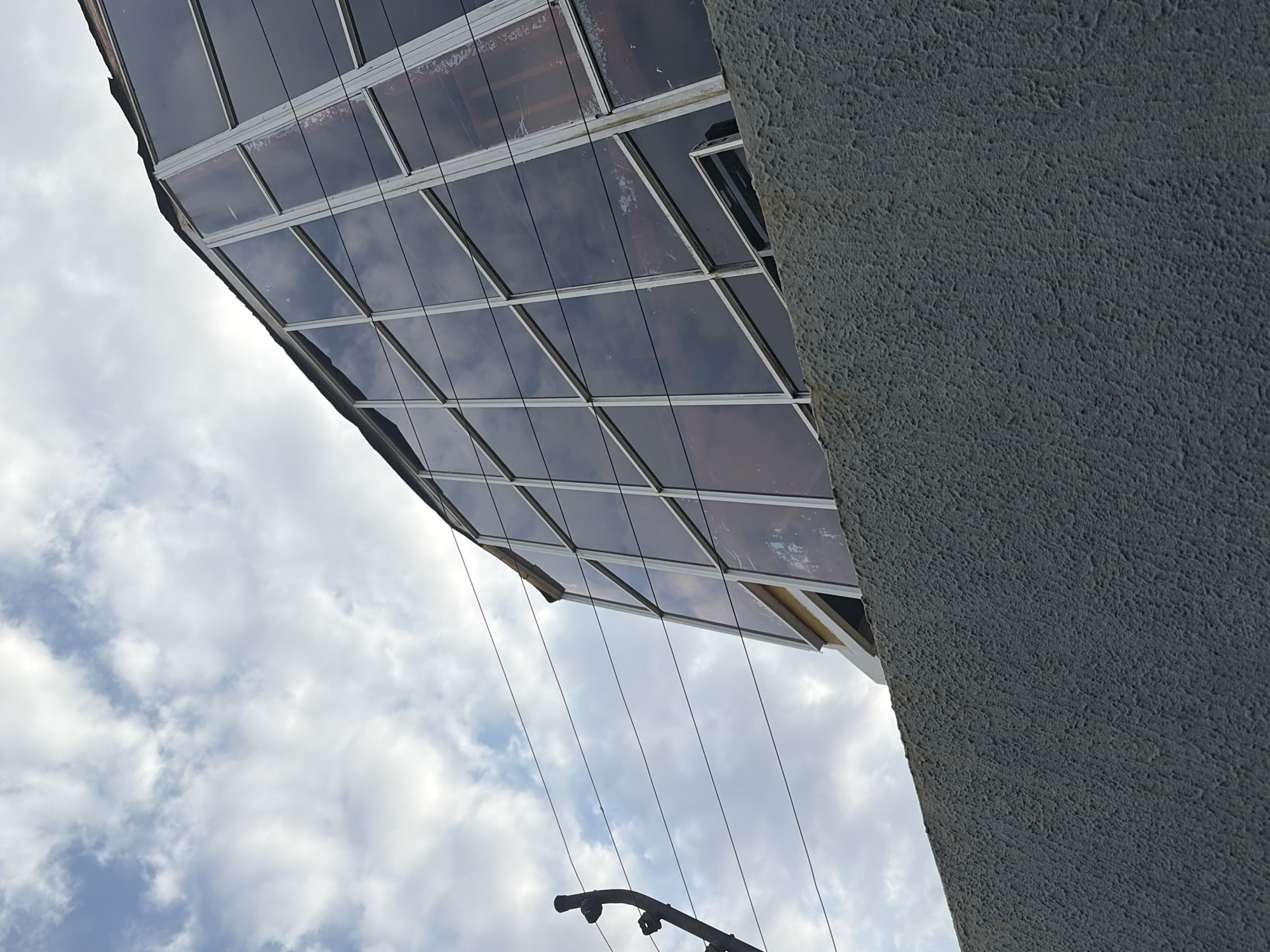 The image depicts an upward view of a building with a grid-like glass facade. The glass reflects the sky, which is filled with white clouds. To the right of the glass facade is a grey concrete wall with a rough texture. At the bottom of the frame, there's a partially visible metal structure with cylindrical components. The building's location is Ciudad Guayana, Venezuela.