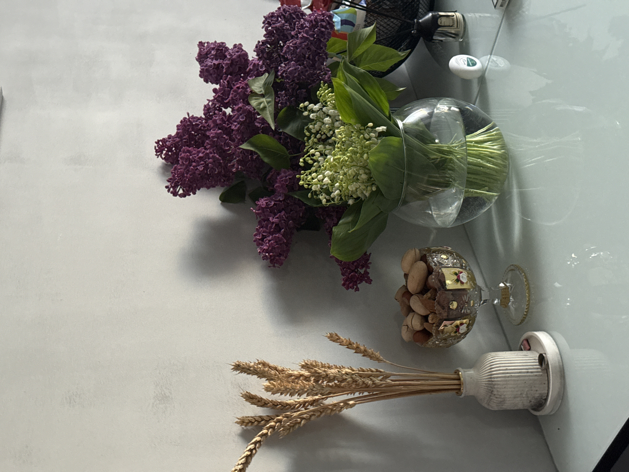 The image displays a still life arrangement on a smooth, light grey or white surface, likely a table or countertop, shot from an overhead perspective. The scene is well-lit, casting soft shadows, suggesting an indoor setting.
The primary subjects are two bouquets of flowers. On the left and center, a large bunch of vibrant purple lilac flowers dominates, showcasing their clustered blossoms and green leaves. To its right, nestled alongside the lilac, is a bouquet of delicate white lily of the valley flowers with their distinctive bell-shaped blooms and broad green leaves. These are arranged in a clear, spherical glass vase containing water, with the green stems visible beneath the water line.
Towards the bottom right of the frame, a decorative, ornate glass goblet filled with various nuts, possibly almonds or similar, stands next to a small, ribbed white ceramic or concrete vase. This vase holds a bundle of dried golden wheat stalks, adding an earthy, rustic element to the composition.
In the upper right background, partially obscured and slightly out of focus, are parts of household items. A black electric fan is visible, with faint text "NU HOME" on a label on its base. Next to the fan, a small, white, oval-shaped object with a green logo and some illegible white text can be seen. Reflections of these objects and the flowers are visible on the glossy surface, indicating a clean and somewhat reflective material. There is no discernible activity or event taking place, as it is a static arrangement of objects. No specific location cues other than an interior setting are present.