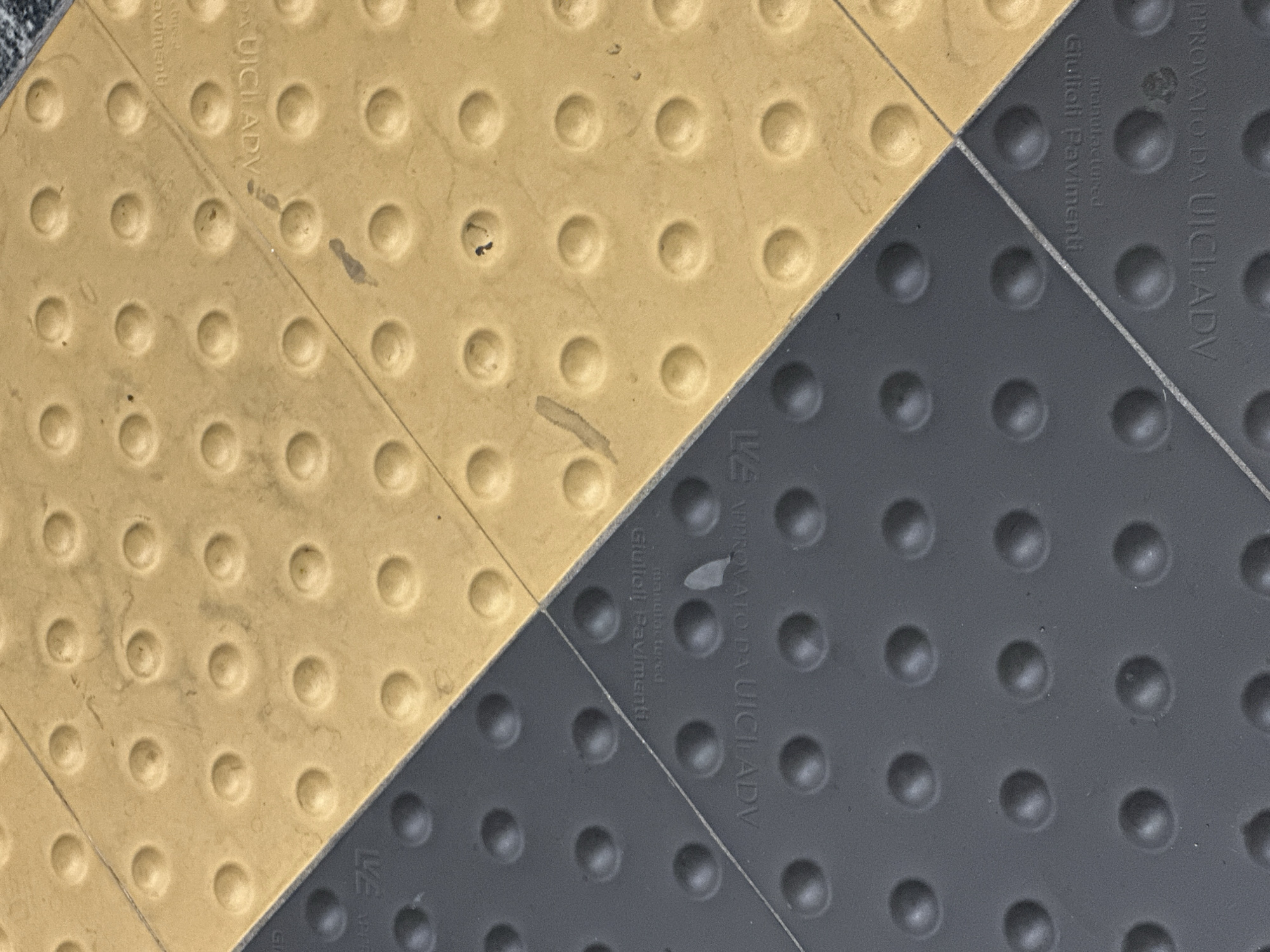 The image shows a close-up, overhead view of two diagonal tactile paving tiles, one yellow and one dark grey, meeting at a seam. Both tiles are square and have a grid of raised, dome-shaped tactile studs. The yellow tile is lightly soiled with a few dark smudges. The dark grey tile also has some faint smudges and a light grey triangular scuff mark. In raised lettering on the yellow tile, partially obscured by the seam and smudges, is text that appears to read "GIULIOTI PAVIMENTI" and "APROVATO DA UICL ADV". Similar text is visible on the dark grey tile. The lighting is even, suggesting an indoor or overcast outdoor environment. The scene is static and appears to be a detail of a pedestrian pathway or public space. Based on the presence of the brand "Giuliotti Pavimenti" and the "UICL ADV" certification, which is often associated with Italian accessibility standards, this paving is likely to be found in a public area in Milan, Italy, such as a street, square, or transit station, designed to assist visually impaired pedestrians.
