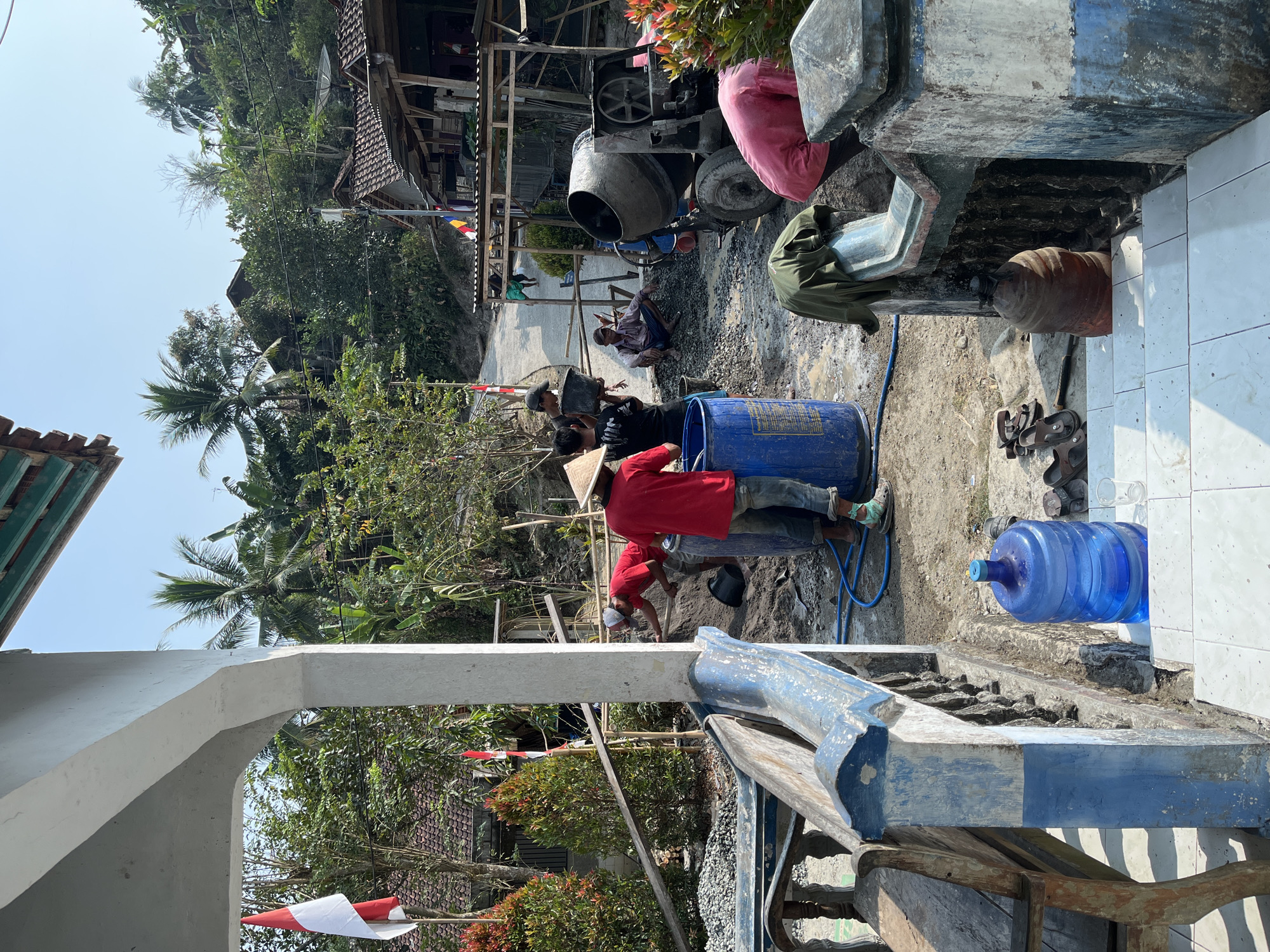 In Kemangguhan, Indonesia, under a clear sky, construction work is underway near a residential area. Several individuals are actively engaged in mixing cement and gravel. One person, wearing a red shirt and a conical hat, is near a large blue drum, possibly preparing the mixture. Another person in a pink shirt is bent over near the cement mixer. A cement mixer stands to the right, ready for use. In the background, traditional houses with tiled roofs are visible, interspersed with lush vegetation and palm trees. Two Indonesian flags are displayed prominently, one on a pole near the building entrance and another smaller one visible in the distance. A wooden bench is placed in the foreground, with a large water dispenser and a glass nearby, suggesting a break area. The ground is a mix of dirt, gravel, and some puddles of water, indicating ongoing construction.
