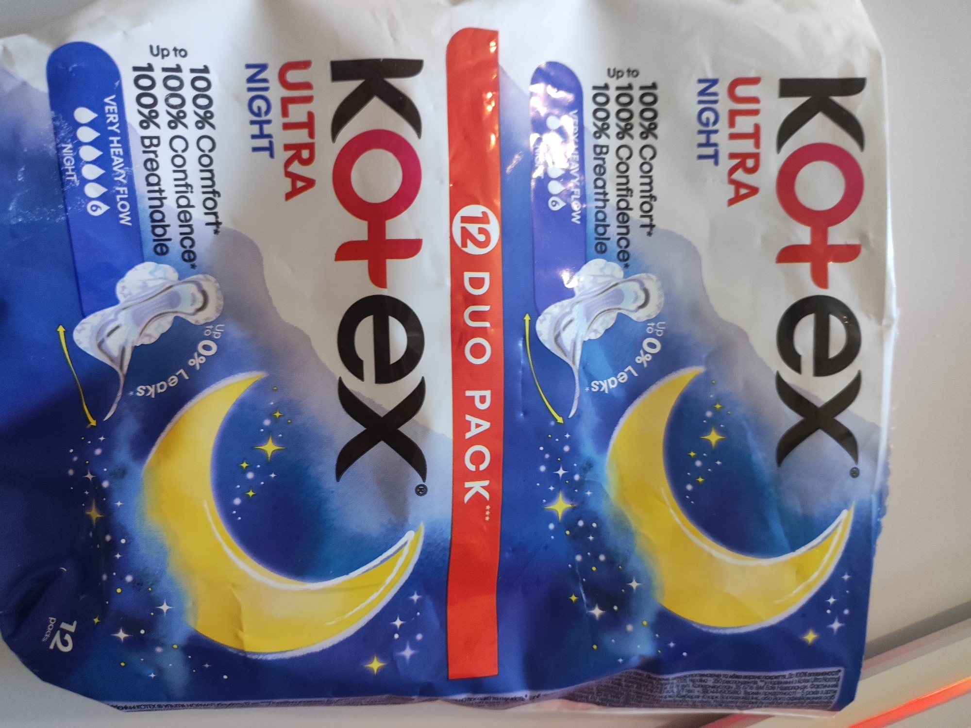 The image shows a package of Kotex Ultra Night pads, with a depiction of a crescent moon and stars on a blue background. The package is partially unwrapped, revealing two individual pads. Text on the packaging indicates "100% Comfort", "100% Confidence", and "100% Breathable". It also states "VERY HEAVY FLOW" and "NIGHT". The number "12" and "pads" are visible on the lower left corner. There is a red vertical band on the package with "12 DUO PACK" written in white. The lighting suggests it is daytime, and the background is plain and white, indicating an indoor setting, possibly a store or home. The image is a close-up of the product packaging. There are no people, animals, or specific landmarks identifiable in the image. The location in Cherkasy, Ukraine, is not discernible from the visual information provided.