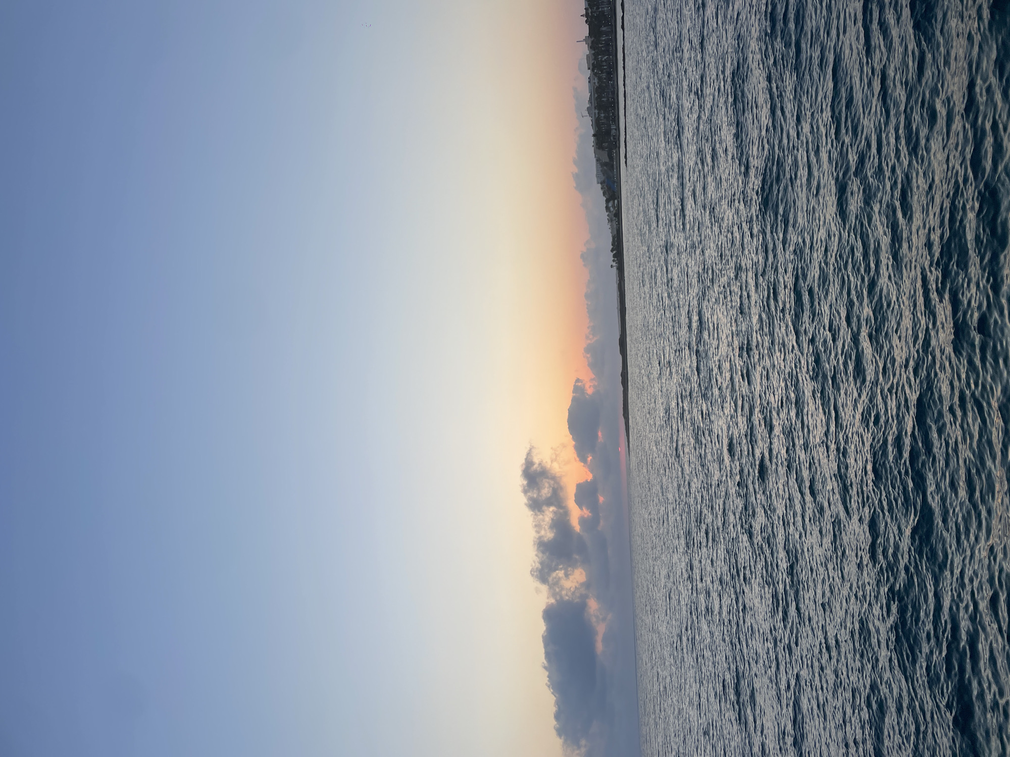 The image depicts a serene coastal scene at dusk, likely in Yeroskipou, Cyprus. The sky transitions from a pale blue at the top to hues of orange and pink near the horizon, indicating the setting or rising sun. A line of fluffy clouds stretches across the middle ground, with some appearing to catch the warm light of the sun. Below the sky is the calm, dark blue expanse of the sea, with subtle ripples on its surface. In the far distance, along the coastline, a faint silhouette of buildings and palm trees can be discerned, suggesting a developed area. The overall atmosphere is peaceful and tranquil, characteristic of a coastal view during twilight.