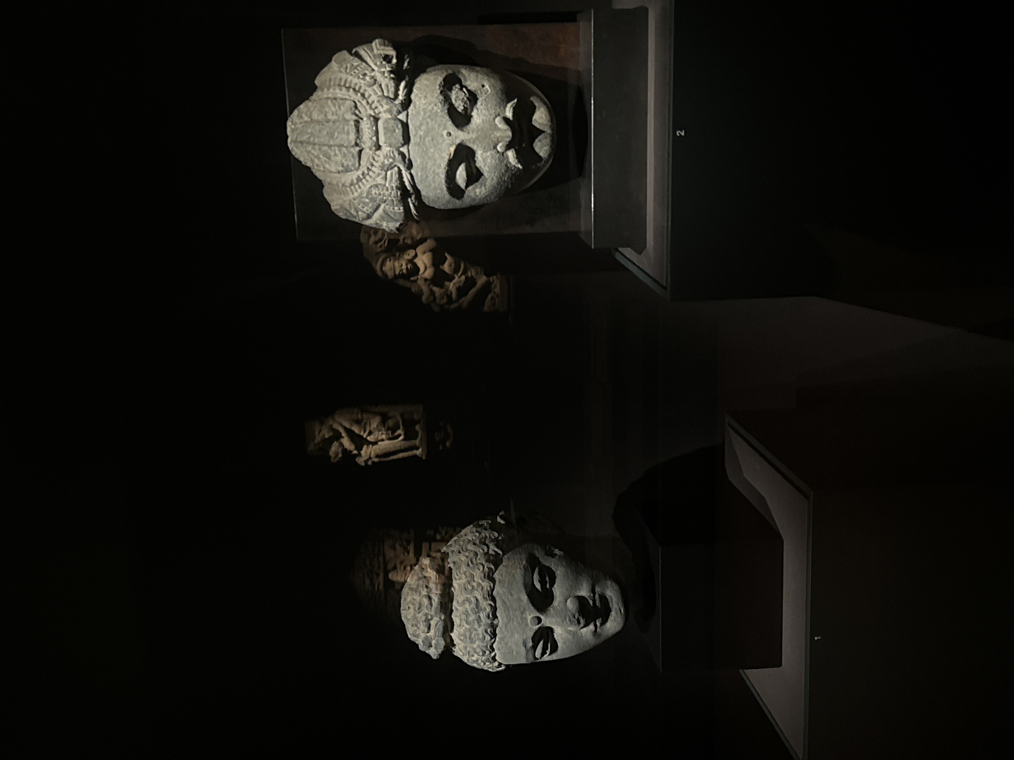 This image displays several stone sculptures in a dimly lit museum exhibition. In the foreground, on the left, is a stone head of a figure with curly hair and serene facial features, placed on a dark pedestal. To the right, a larger stone head with a headdress, mustache, and pronounced facial features is prominently displayed on a dark, rectangular base. In the background, several smaller, less distinct sculptures are visible, including one that appears to depict a human figure in a devotional pose. The overall atmosphere is dark and dramatic, with focused lighting on the exhibited artifacts.
The specific location in Seoul, South Korea, cannot be determined from the provided image.