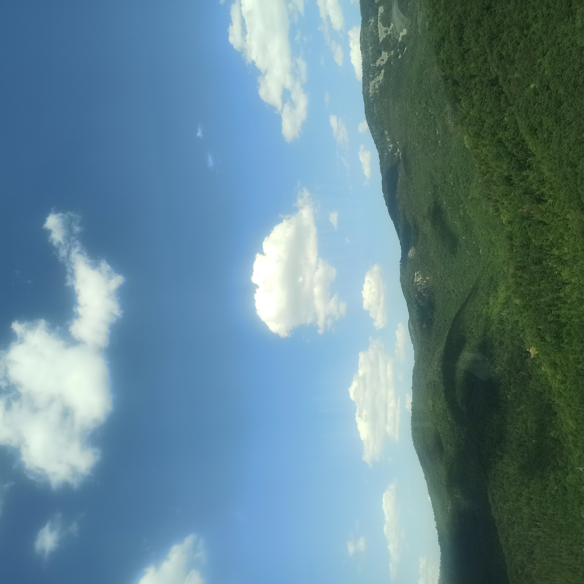 A bright, clear blue sky with scattered white cumulus clouds dominates the upper half of the image. Below the sky, a vast, rolling landscape of lush green mountains is visible. The mountains are densely covered with trees, creating a textured, verdant surface. Shadows are cast across the slopes, indicating the direction of sunlight. In the distance, the peaks of the mountains appear somewhat rugged, with areas of lighter vegetation or rock exposed on some of the ridges. The overall scene depicts a natural, undisturbed environment in the mountains near Vrachesh, Bulgaria. There are no people or man-made structures clearly visible in this landscape image.