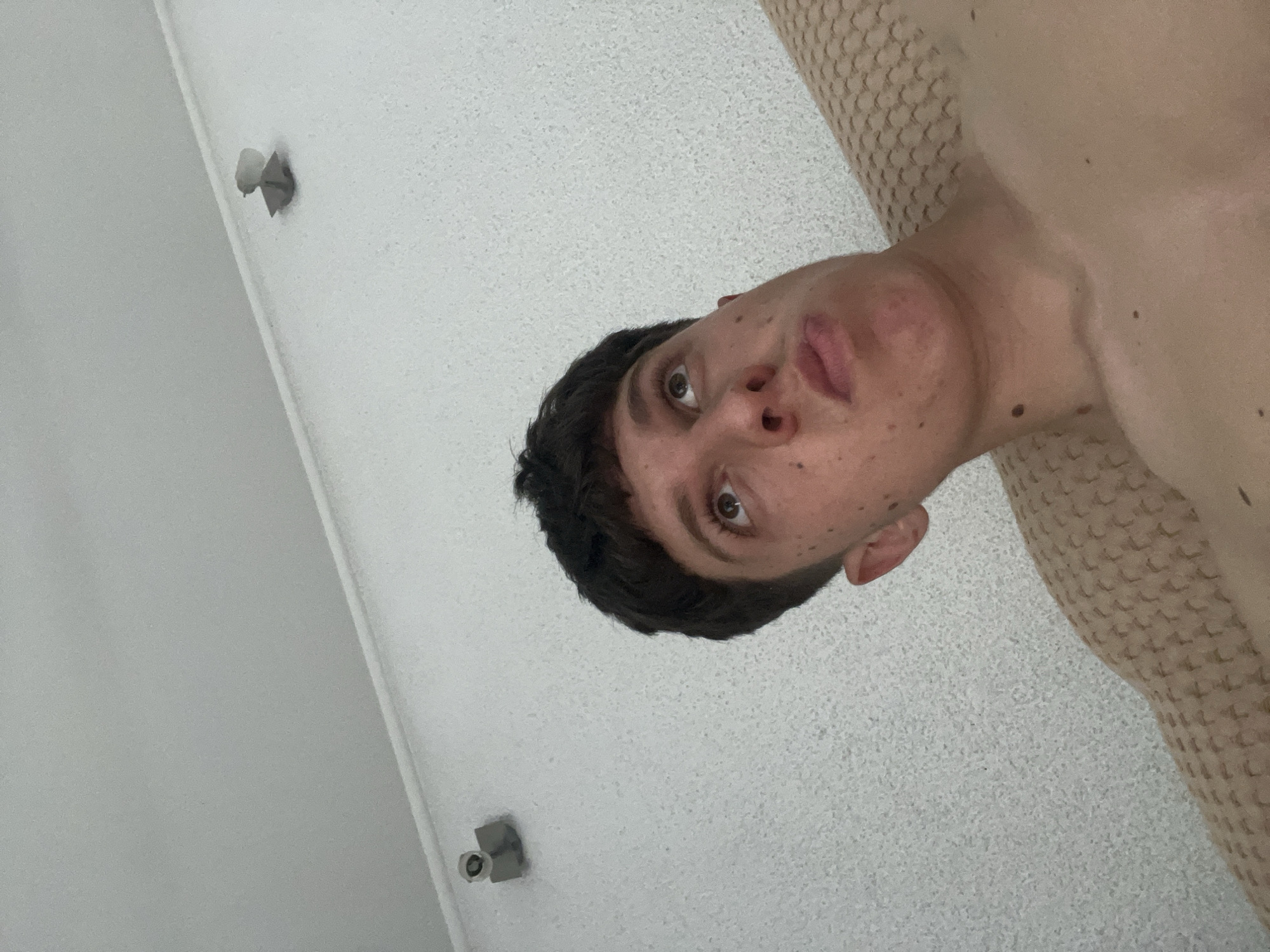 An indoor scene in Zlatograd, Bulgaria, depicts a young male from the mid-chest upwards, observed from a low angle looking upwards. The individual has short, dark hair styled in a bowl cut and several small moles on his face and upper body. His gaze is directed upwards and slightly to his left, and his lips are pursed. He appears shirtless or minimally clothed, and a textured, light-brown fabric, possibly a blanket or furniture upholstery, is visible on the right side of the frame adjacent to his shoulder. The background features a light-colored wall with a pronounced stucco-like texture. Two small, cylindrical security cameras or sensors are mounted on this textured wall, one clearly visible in the upper left quadrant and another partially visible in the upper right. Above the textured wall, a smooth, light-colored ceiling is visible, separated by a crisp horizontal line.