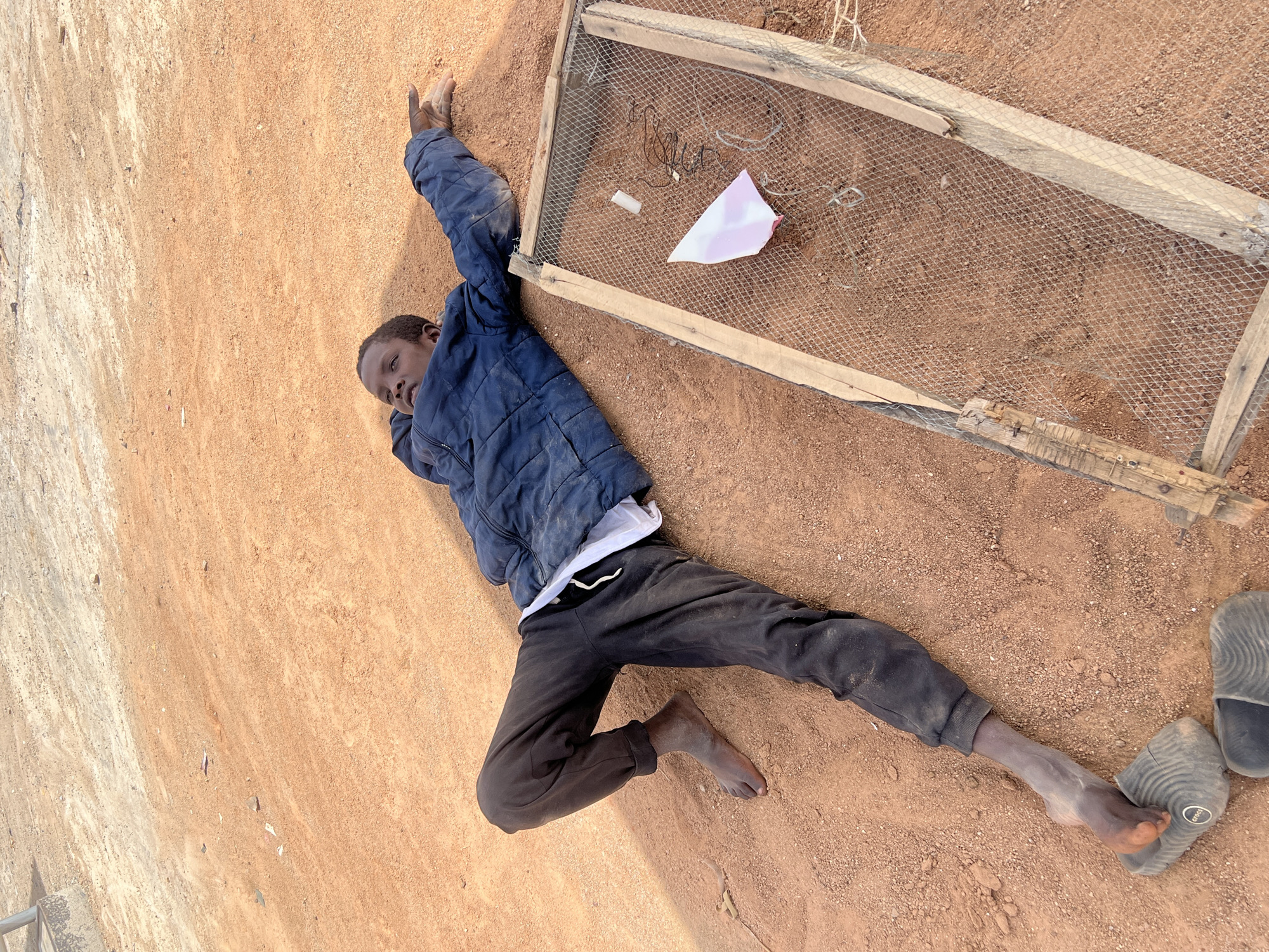 A man lies on the ground in Barakin Lerawi, Nigeria. He wears a blue jacket, black pants, and a white undershirt. His feet are bare except for one sandal near his right foot. A rectangular wooden frame with wire mesh sits to his right. A small, torn piece of pink paper and some wires are on the mesh surface. The ground appears to be a mixture of sand and dirt, with varying shades of brown and tan. The man's left arm is extended above his head.