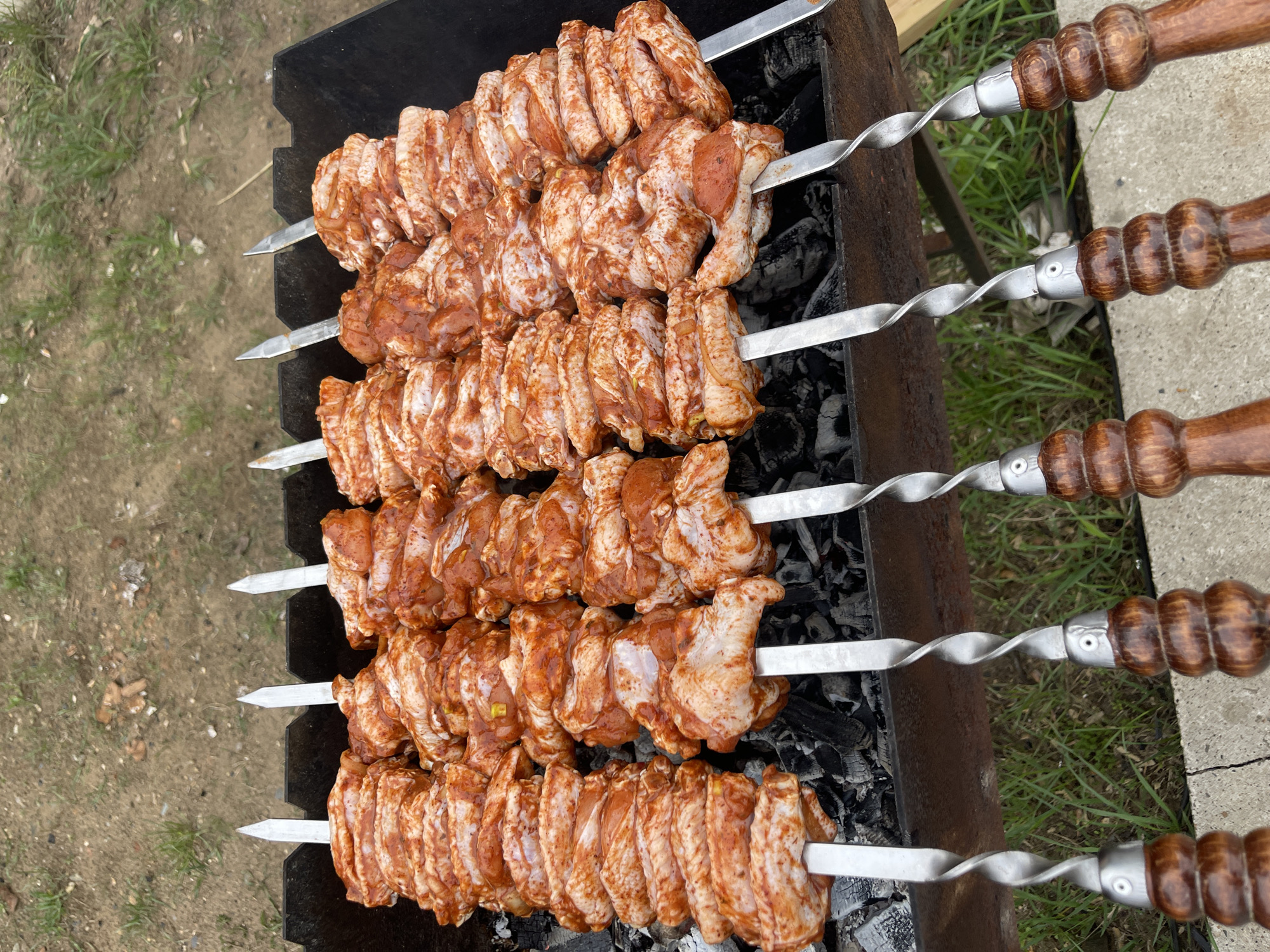 The image shows chicken wings, marinated in a red-tinged sauce, arranged on five metal skewers over a charcoal grill. The grill is positioned outdoors, likely in a backyard or similar setting. The skewers have wooden handles. The location is Chaikovsky, Russia.