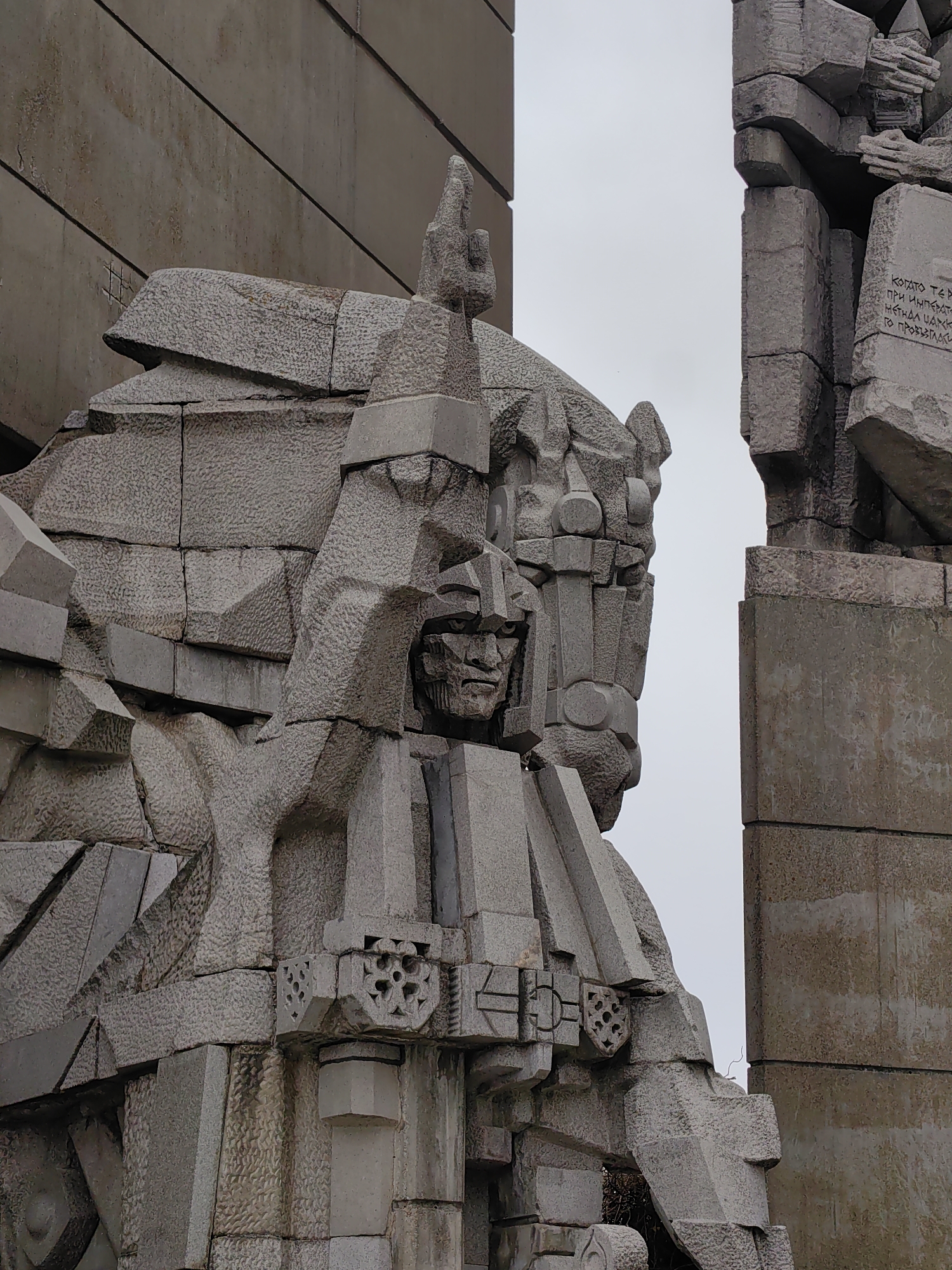 The image shows a close-up of a large, abstract, gray stone monument. The monument is composed of large, roughly hewn blocks, forming a figure with a helmeted face and a stern expression. Intricate carvings and geometric patterns decorate the monument. The sky is overcast and white. The monument is part of the Founders of the Bulgarian State monument complex, located on the outskirts of Shumen, Bulgaria. The monument commemorates the 1300th anniversary of the Bulgarian state.