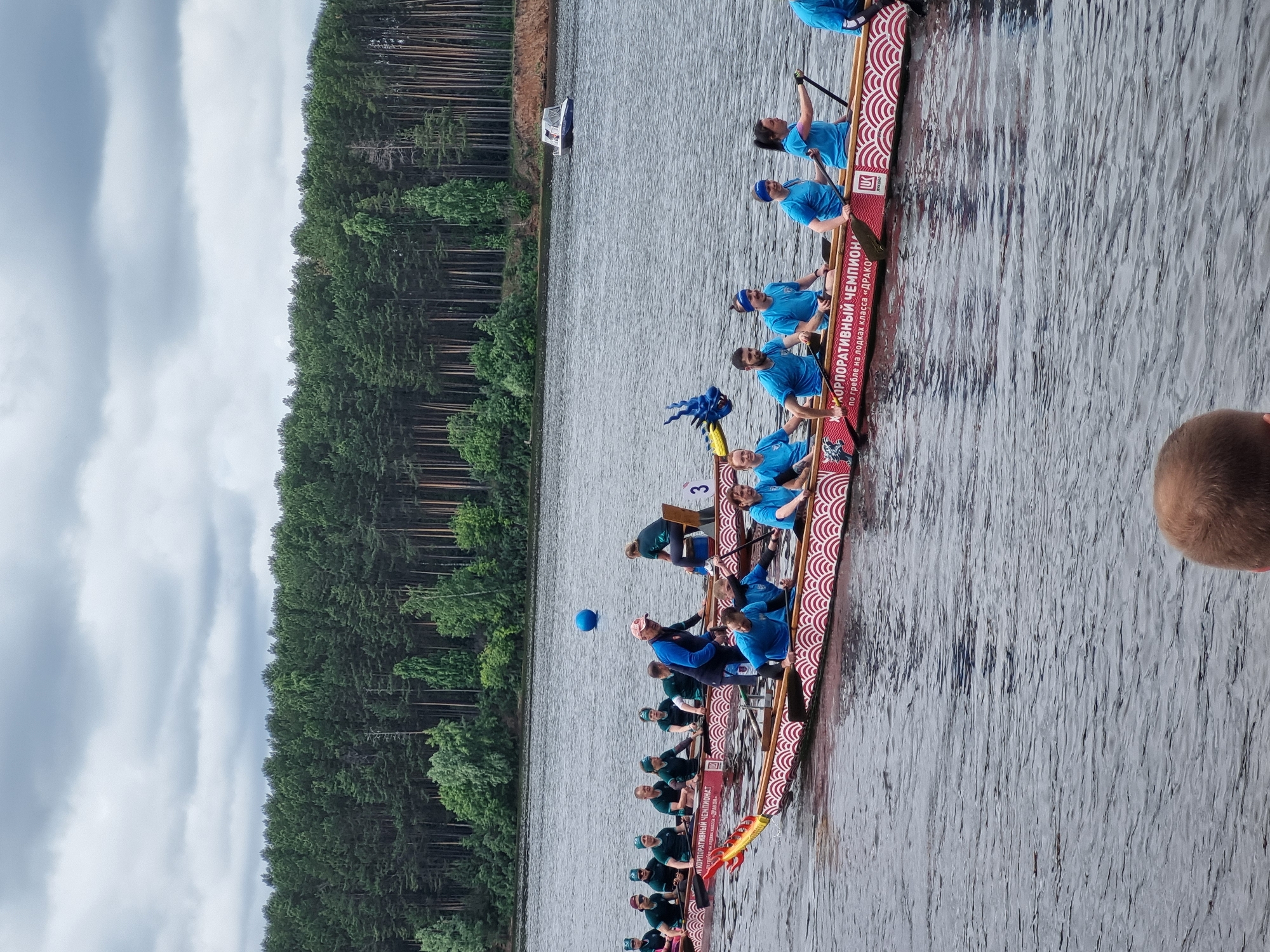 An overcast day at the Kama River in Chaikovsky, Russia, hosts a dragon boat race. The foreground is dominated by a long, narrow dragon boat adorned with red and white wave patterns, featuring a yellow and red dragon head at the bow and a blue dragon crest at the stern. Ten individuals, dressed in blue t-shirts and headbands, are actively paddling the boat. Their faces show exertion and focus as they synchronize their strokes. Behind this boat, another dragon boat is partially visible, also with paddlers. In the distance, a dense, dark green forest lines the riverbank under a cloudy sky. A small white boat is moored near the shore. The water is a deep grey, reflecting the subdued lighting. The overall atmosphere is one of intense athletic competition within a natural setting.