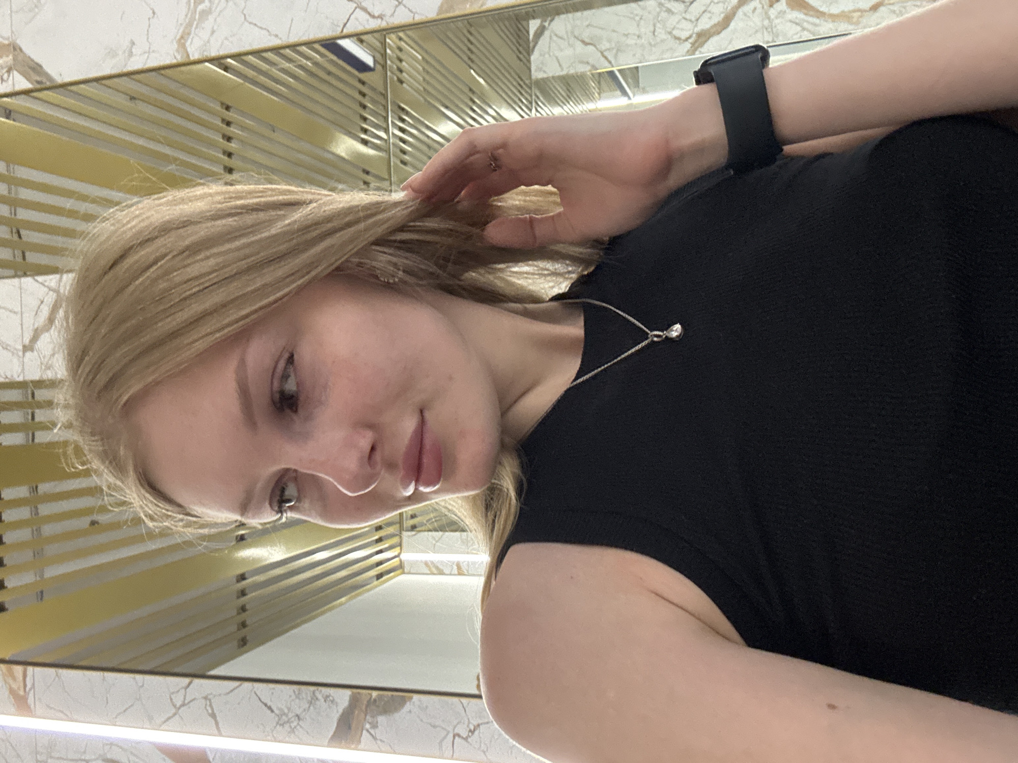 A young woman with blonde hair is taking a selfie in what appears to be a modern interior, possibly a hotel lobby or a high-end establishment. The background features a mirrored surface reflecting vertical golden and white striped elements, suggesting a contemporary design with metallic accents. The woman is wearing a black sleeveless top and a silver necklace with a pendant. Her left arm is extended, with her hand touching her hair, and she is wearing a black smartwatch on her right wrist. The image is taken from the chest up, with the woman looking to her left. There are no discernible landmarks or contextual clues within the image that would specifically place it in Perm, Russia.