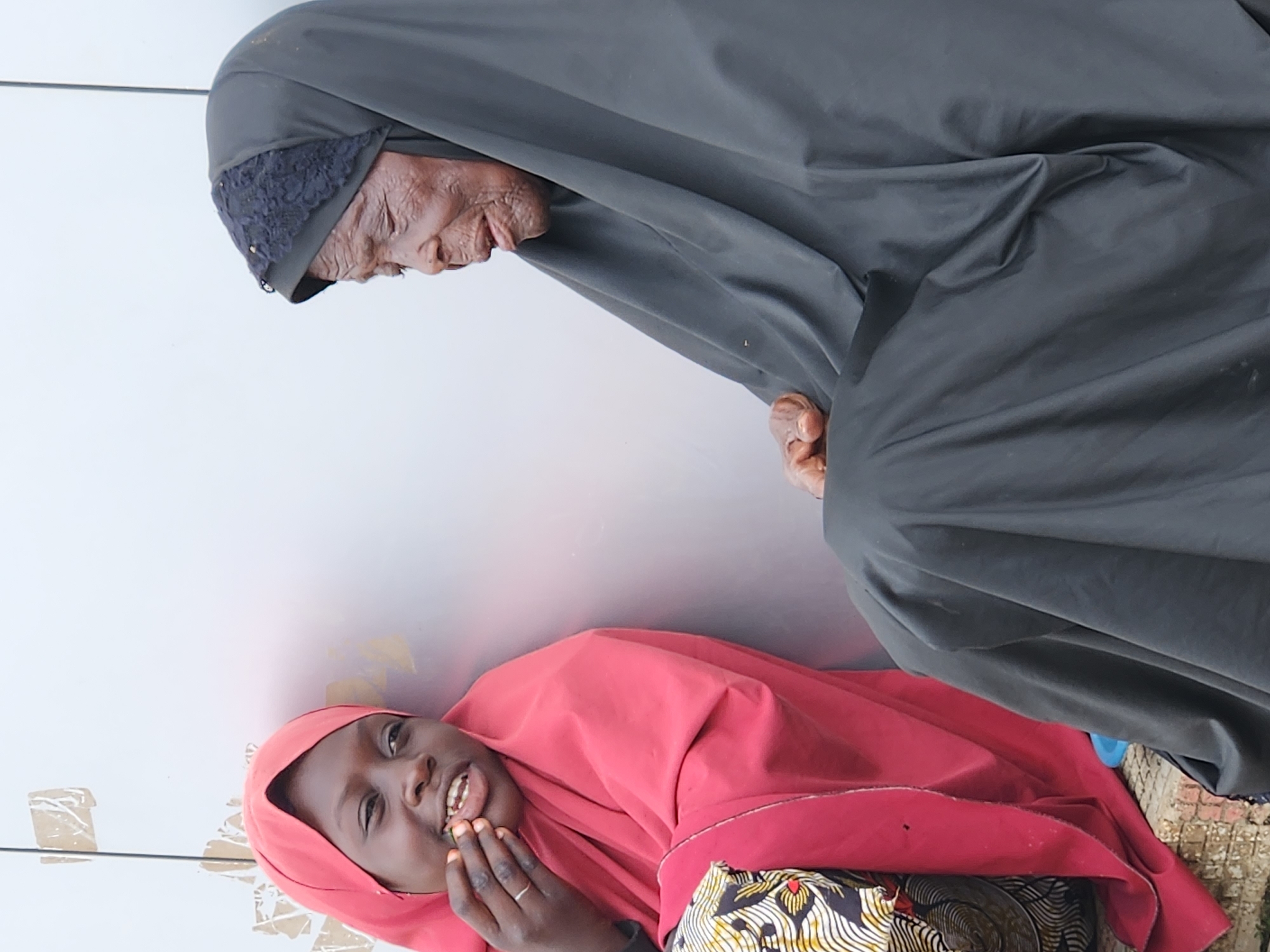 Two individuals, a younger female and an older female, are sitting next to each other outdoors. The younger female, on the left, is wearing a pink hijab and a colorful patterned dress. She is smiling and touching her mouth with her hand. The older female, on the right, is wearing a dark grey hijab and is looking to the side with her eyes closed. They are seated on what appears to be a low wall or step. The background is a plain, light-colored wall with some scuff marks.