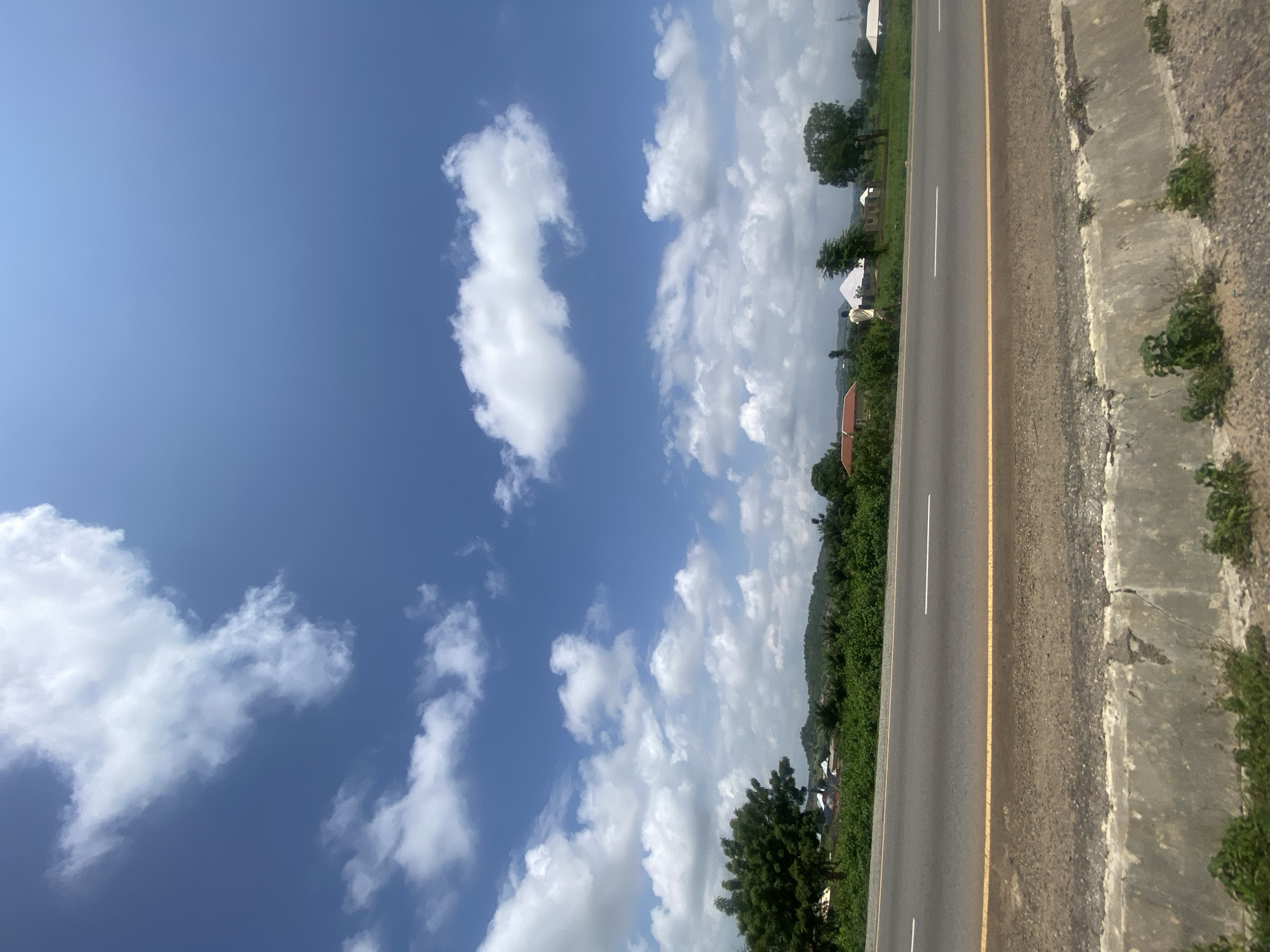 The image depicts a road scene in Sobon-Gida, Nigeria, under a bright blue sky with scattered white clouds. The road, paved with asphalt, is marked with a solid yellow line along the right edge and a dashed white line in the center, indicating two lanes of traffic. A concrete barrier runs along the right side of the road, with vegetation growing on its edge. Beyond the road, lush green trees and dense foliage form a natural landscape, interspersed with a few buildings, some with red-tiled roofs and others with white roofs. In the distance, rolling hills create a layered horizon. A lone figure, a male wearing a light-colored garment, is visible walking along the edge of the road amidst the greenery and buildings. The overall scene suggests a rural or semi-urban environment in Nigeria.
