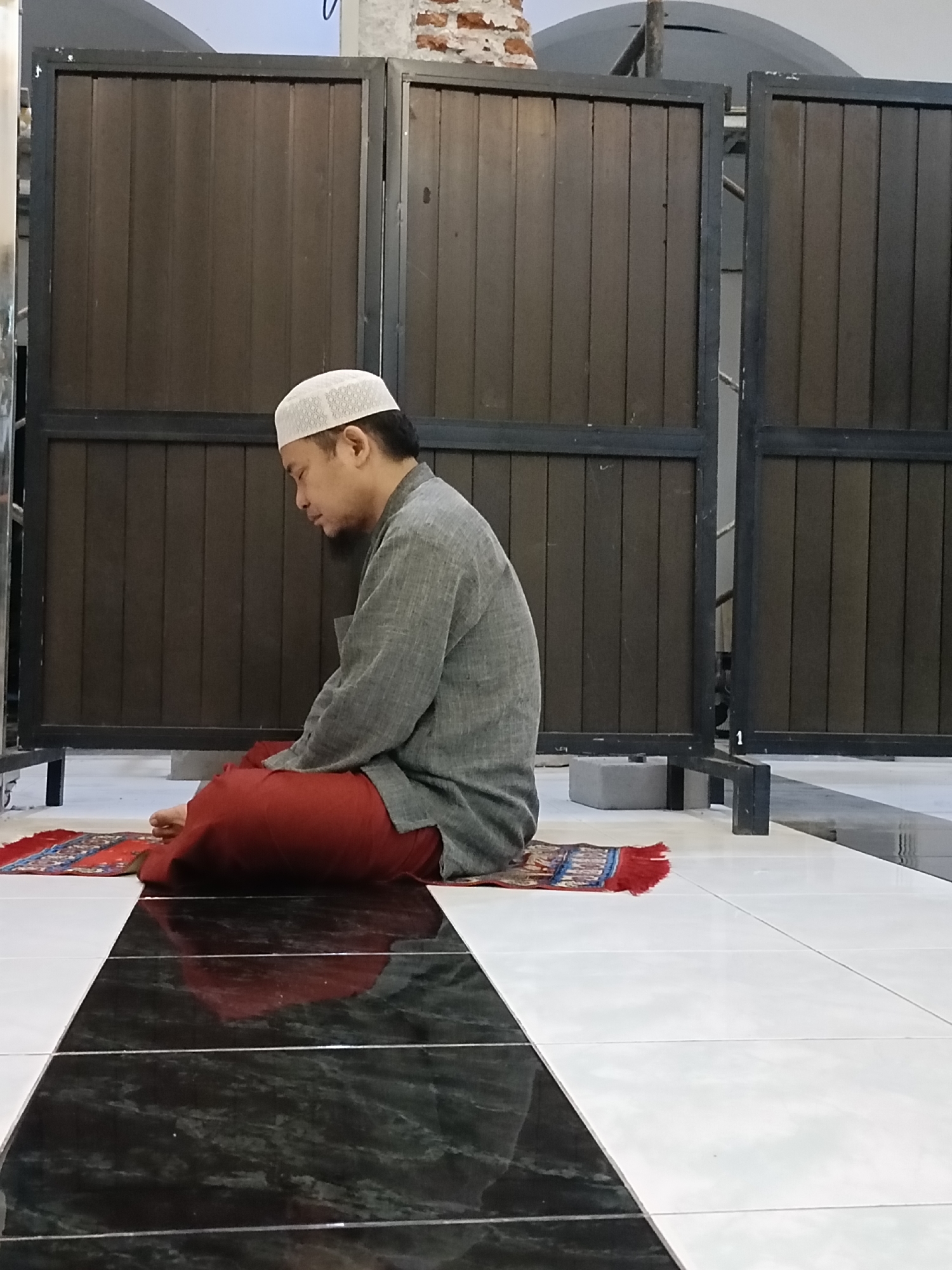 A man wearing a white skullcap and a grey robe is sitting on a prayer mat in a room. The man is facing left and appears to be praying. The prayer mat is red and has a pattern on it. The room has a tiled floor with black and white tiles. Behind the man are two dark brown wooden panels. The background also shows some exposed brick and a curved ceiling. The scene appears to be a prayer room, likely in a mosque or a private residence, located in Kediri City, Indonesia.