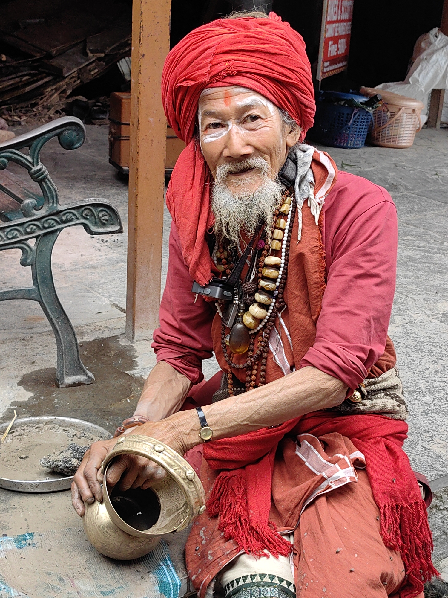 In Vashisht, India, a weathered man with a white beard and red turban, adorned with white face paint, sits with a brass pot in his hands. He wears traditional Indian clothing, including a red scarf with fringe, and multiple beaded necklaces. A dark green metal bench and a weathered wooden pole are visible in the background, along with a metal tray containing sand and a rock. The ground is paved with grey stones.