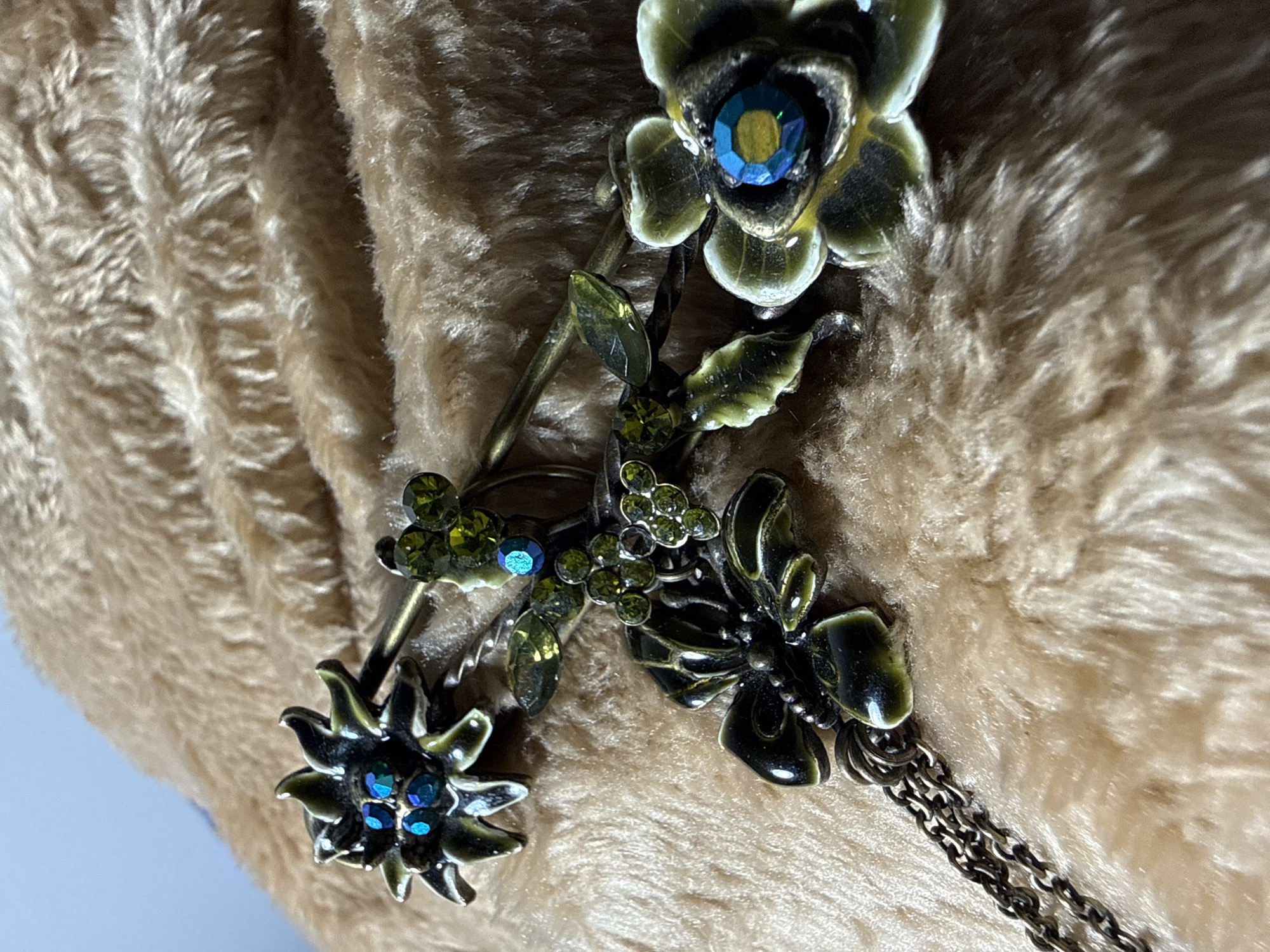 A close-up shot displays a vintage-style flower and dragonfly pendant lying atop a soft, plush surface. The pendant features intricate metalwork, with painted green and white floral and insect motifs accented by small, sparkling blue and green rhinestones. A delicate chain extends from the dragonfly component. The plush surface beneath the pendant is a light brown, adding warmth to the shot. There is no indication of this picture being taken in Moscow, Russia.