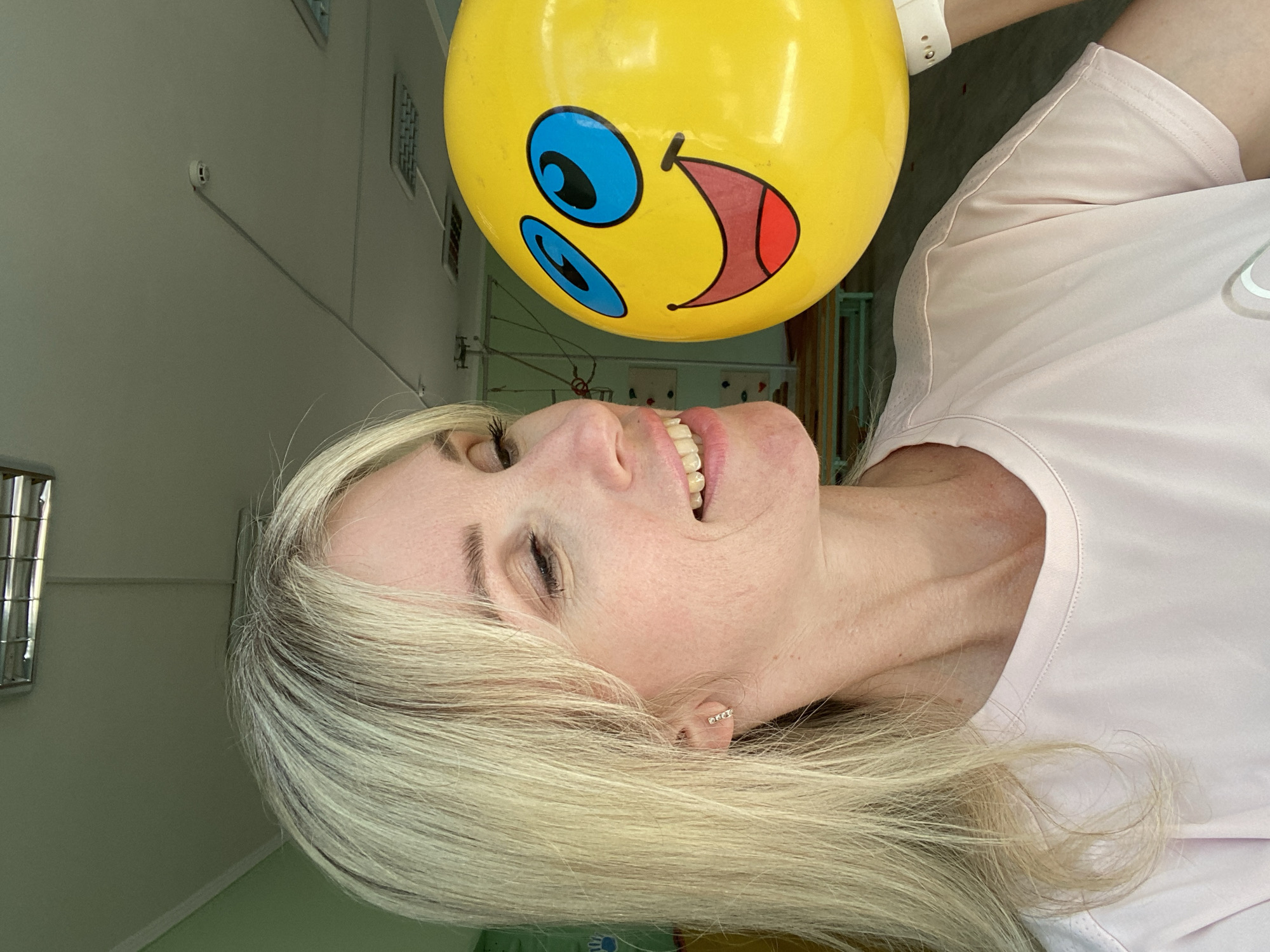 A medium shot shows a fair-skinned woman with blonde hair and bangs, wearing a light pink t-shirt, looking at a large yellow balloon with a smiley face. The woman is positioned on the left side of the frame, her head tilted as she gazes at the balloon. The smiley face on the balloon features large blue eyes, black pupils, and a wide, red-lined mouth with black lips. The balloon is held by the woman's right hand, which is partially visible on the right edge of the frame, wearing a white watch. The background appears to be an indoor space with a light-colored ceiling, visible light fixtures, and some green walls. There are also some blurred elements in the background that suggest a room with equipment, possibly a classroom or play area, with a wooden bench visible. The scene is brightly lit.
