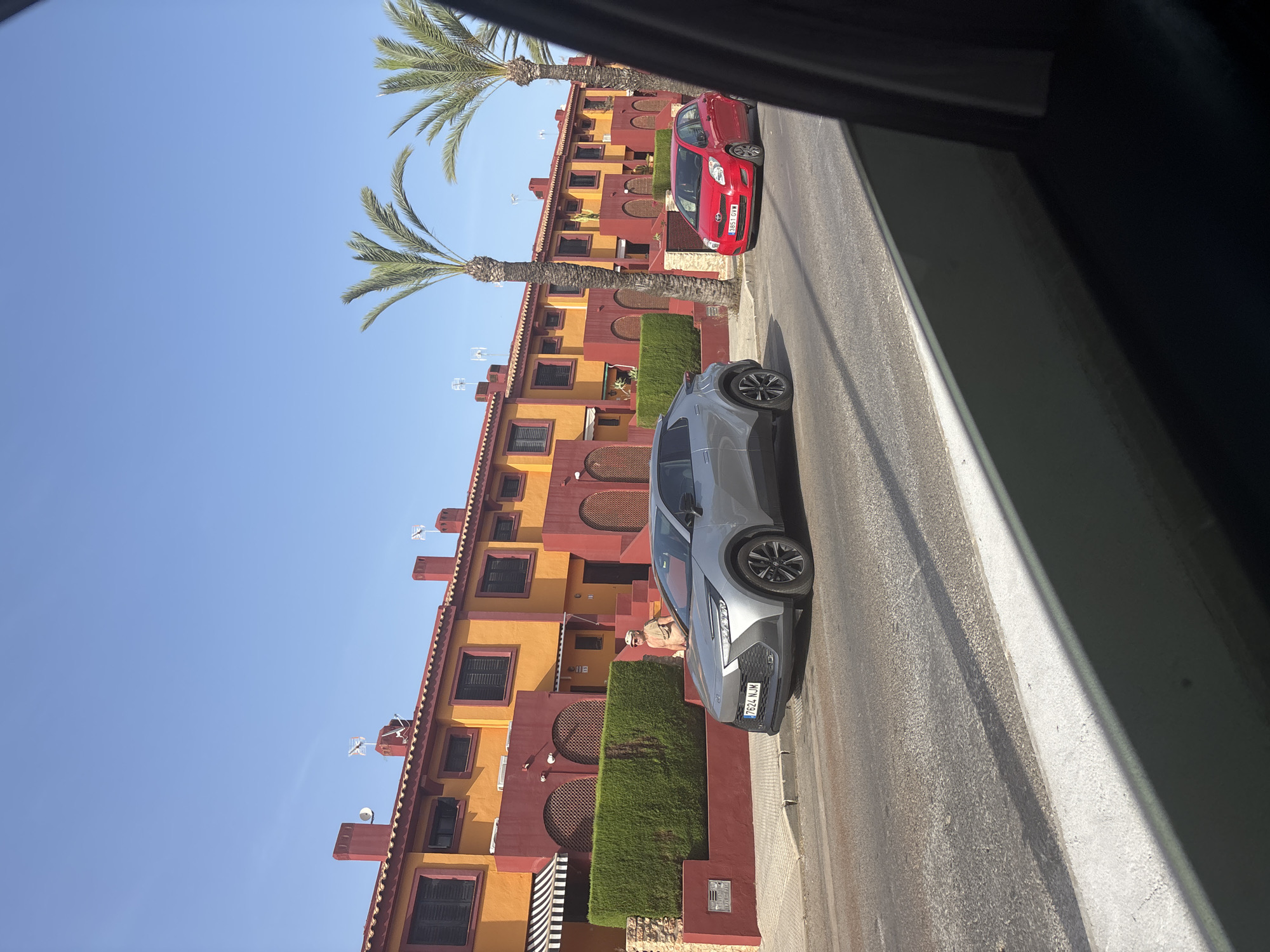 A street scene in an urban area features a row of attached houses with orange and red facades under a clear blue sky. Two cars are parked on the side of the road in front of the houses: a silver hatchback on the left and a red hatchback on the right. A person in a light-colored shirt and hat stands near a green hedge in front of one of the houses. Palm trees are visible in the background. The foreground shows a portion of a road with a white curb and a dark shadow.