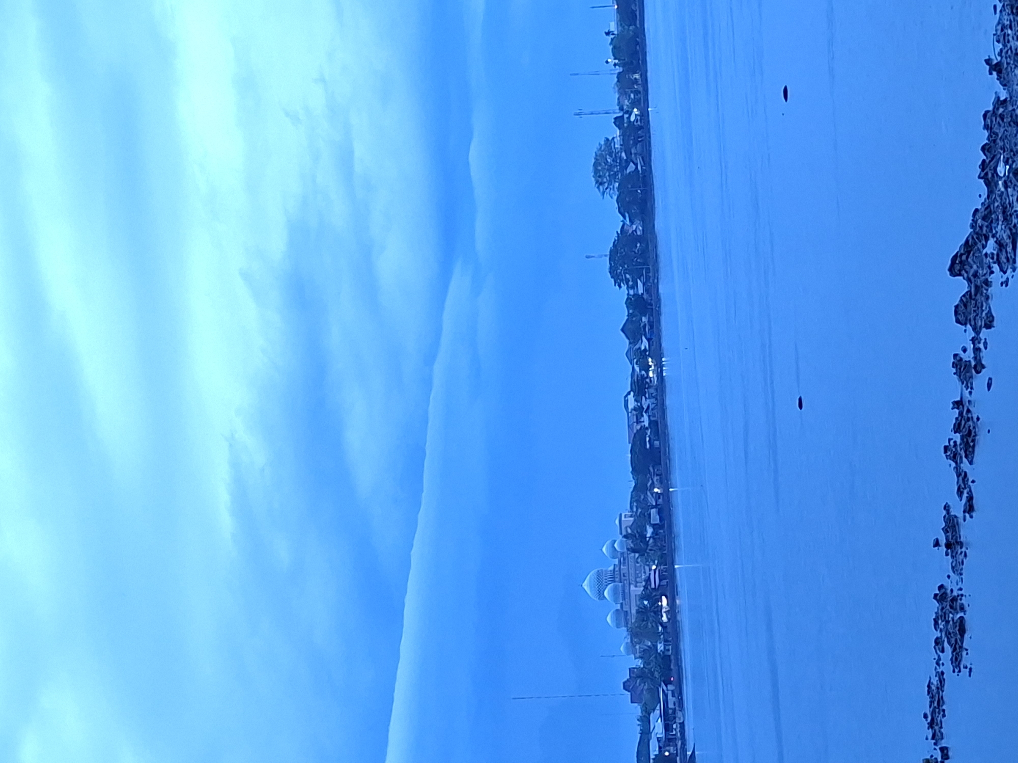 This serene landscape captures a wide expanse of water under a vast, soft blue sky, likely at dawn or dusk, or on an overcast day, dominated by cool blue and cyan hues.
The sky, filling the upper portion of the frame, is a blend of light blue and cyan, featuring subtle, diffused cloud formations. A distinctive, long, horizontal cloud band stretches across the upper-middle section, adding a linear element to the sky.
Below the horizon, an expansive body of calm water, possibly a sea or large river, stretches towards the foreground. Its surface is smooth and reflects the sky's color. In the far distance on the water, a small, dark, indistinct object can be seen. Along the distant left shoreline, a city or townscape is visible, characterized by a line of buildings and trees. Prominently featured among these structures is a large building complex with multiple domes and spires, strongly indicative of a mosque. Several small, bright lights dot the distant shore, suggesting either streetlights or interior lights of buildings. Towards the bottom-right, a darker, textured mass resembling rocky formations or a rugged coastline extends into the water.
The scene conveys a sense of tranquility and vastness, with the distant urban elements adding a subtle human presence to the natural setting. No specific activity is discernible, and no text is visible. The architecture of the mosque aligns with the provided location context of Lhokseumawe, Indonesia.