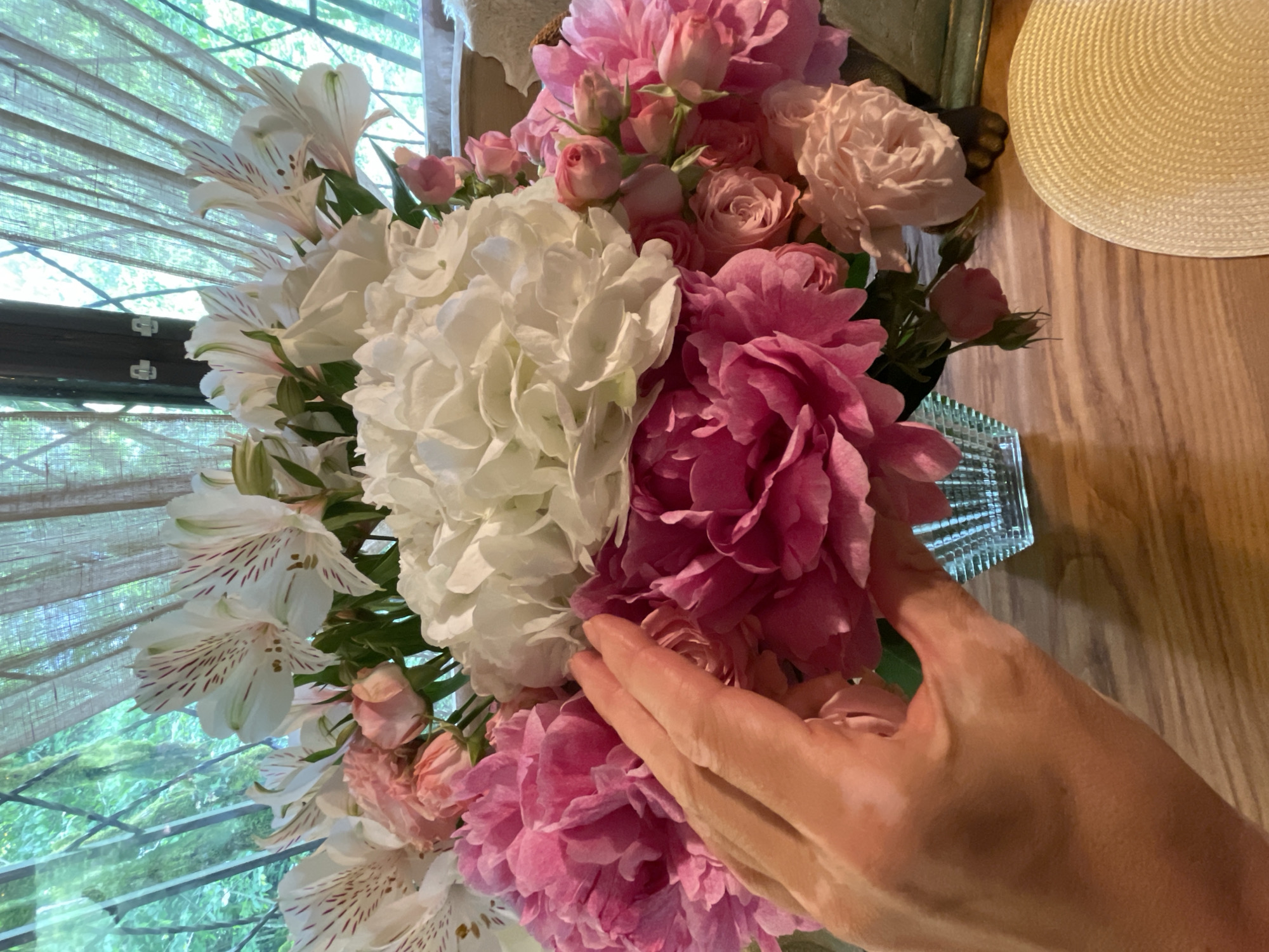 A high-angle medium shot depicts a bouquet of flowers and a hand, possibly indoors, in what could be Solokh-Aul, Russia. The bouquet features a mix of white alstroemeria, white hydrangea, pink peonies, and pink roses. A hand is touching the pink peonies. The flowers are arranged in a clear glass vase. A straw hat sits on a wooden surface in the upper right corner of the frame. A window with blinds is visible in the background.