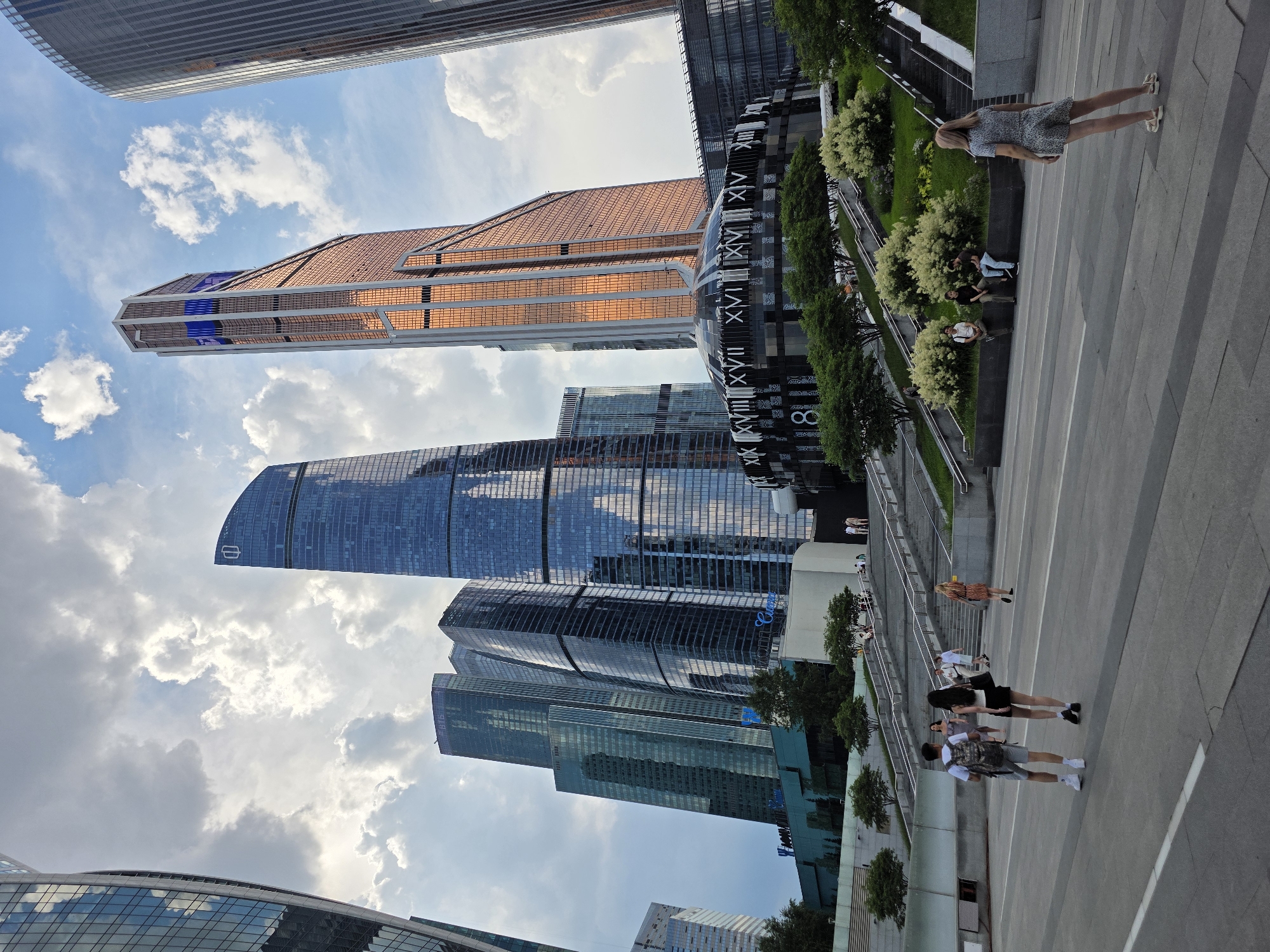 This image depicts a view of the Moscow International Business Center, commonly known as Moscow City, in Moscow, Russia. The scene is dominated by several modern skyscrapers with glass facades reflecting the sky and clouds. In the foreground, there is a wide paved area with several people walking and standing.
In the mid-ground, a circular building with Roman numerals on its exterior is visible, situated at the base of a copper-colored skyscraper. A large set of stairs leads up to a tiered garden area with trees and bushes, where more people are seated or walking. The sky is partly cloudy with a mix of blue sky and white clouds.
Specific details include:
- **Architecture:** Numerous skyscrapers of varying heights and designs, including glass-paneled towers and the distinctive copper-colored skyscraper with blue accents. The circular building features Roman numerals, suggesting a clock or artistic element.
- **People:** Several individuals are scattered throughout the scene. A couple is walking towards the stairs in the foreground, a woman stands facing away from the camera on the right, and groups of people are seated on a ledge near the gardens.
- **Environment:** The scene is set in an urban plaza with a modern aesthetic. Landscaping with trees and bushes softens the concrete and glass structures. The paving stones in the foreground are laid in a patterned manner.
- **Time of Day:** The lighting suggests it is late afternoon or early evening, with shadows beginning to lengthen.
- **Action:** People are engaged in walking, standing, and sitting, indicating a public space being utilized for leisure and transit.