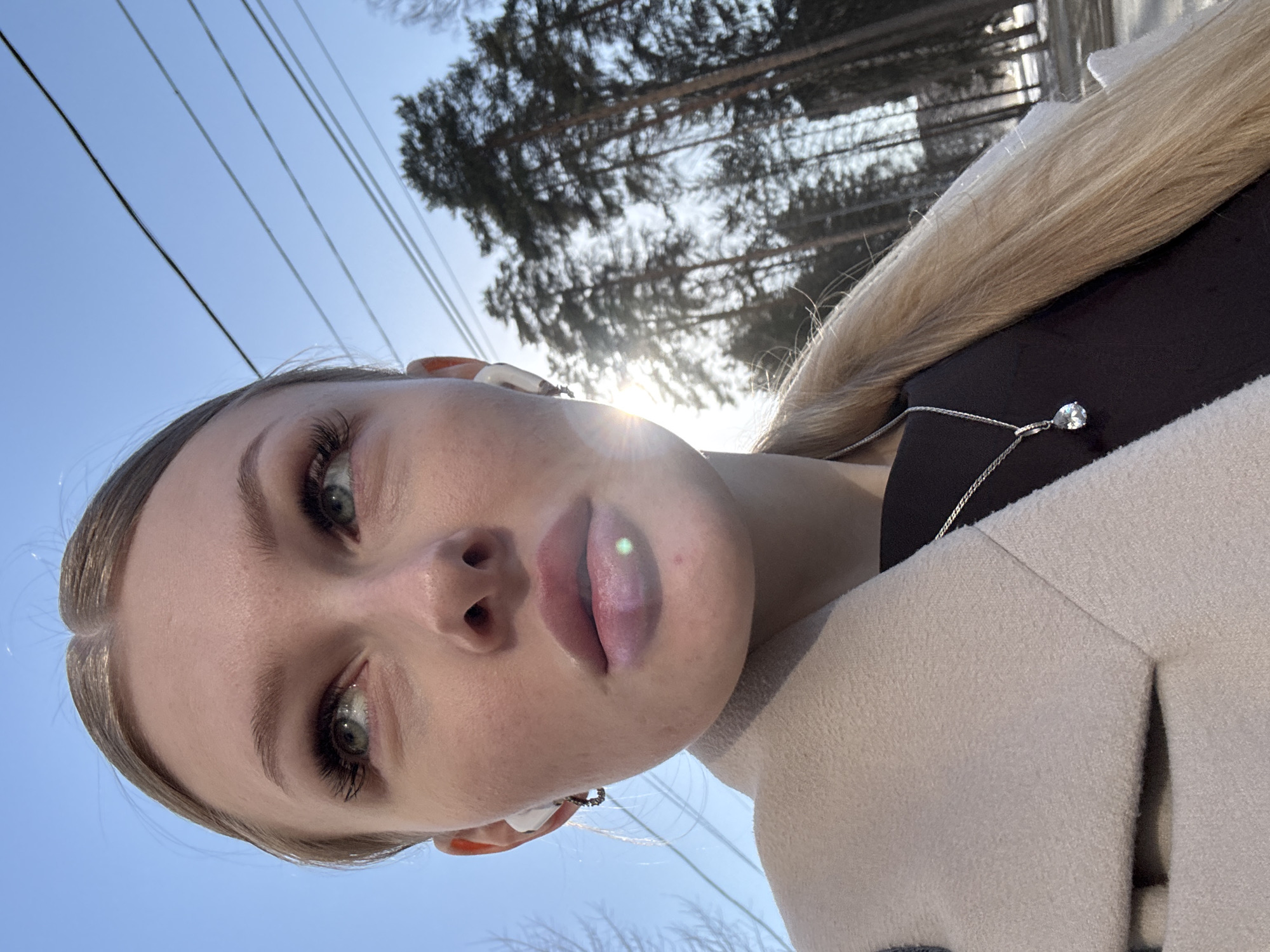 A young woman with blond hair, green eyes, and fair skin takes a selfie outdoors on a sunny day. She is wearing a light-colored coat and a necklace with a teardrop pendant. Her lips are slightly parted. In the background, tall trees are silhouetted against the bright sun and clear blue sky. Overhead power lines are visible. While it is not possible to confirm this image was taken in the city of Perm, Russia using visual information alone, the landscape resembles that of the region.
