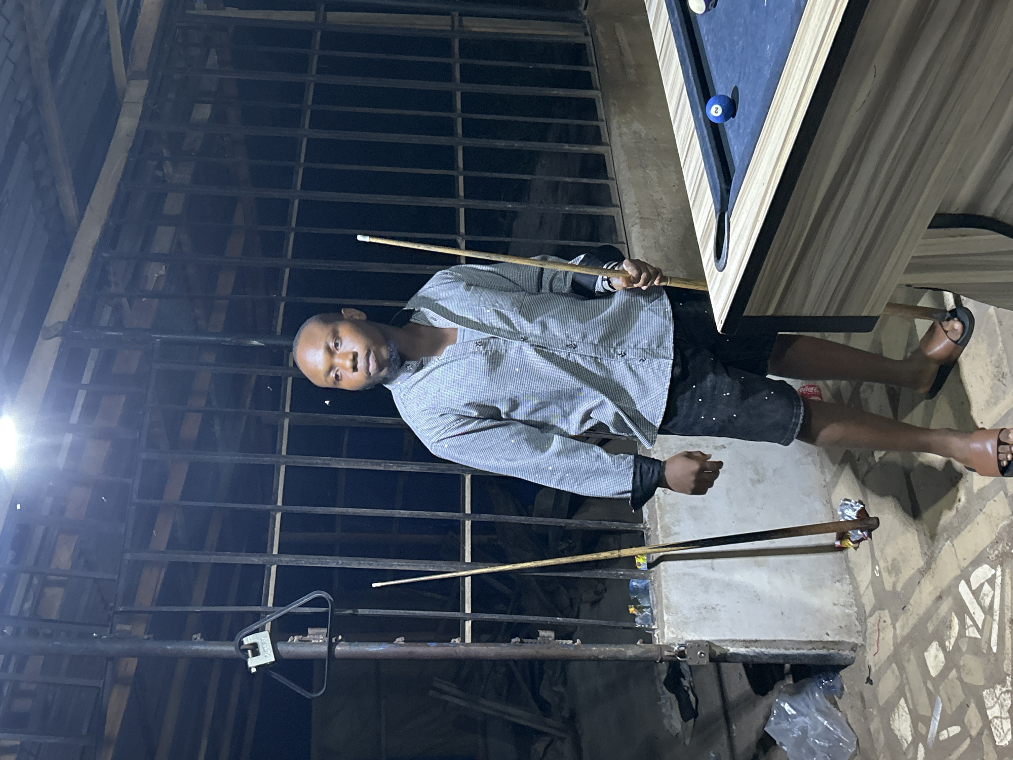 A man is standing next to a pool table outdoors in Ihiagwa, Nigeria. He is holding a pool cue in his right hand and has another cue resting on a nearby structure. The pool table has at least one ball on it. The man is wearing a grey shirt and dark shorts, and is wearing sandals. The location has a metal latticework structure in the background, and the ground appears to be paved with patterned bricks or tiles. Debris is on the ground near the bottom of the frame.