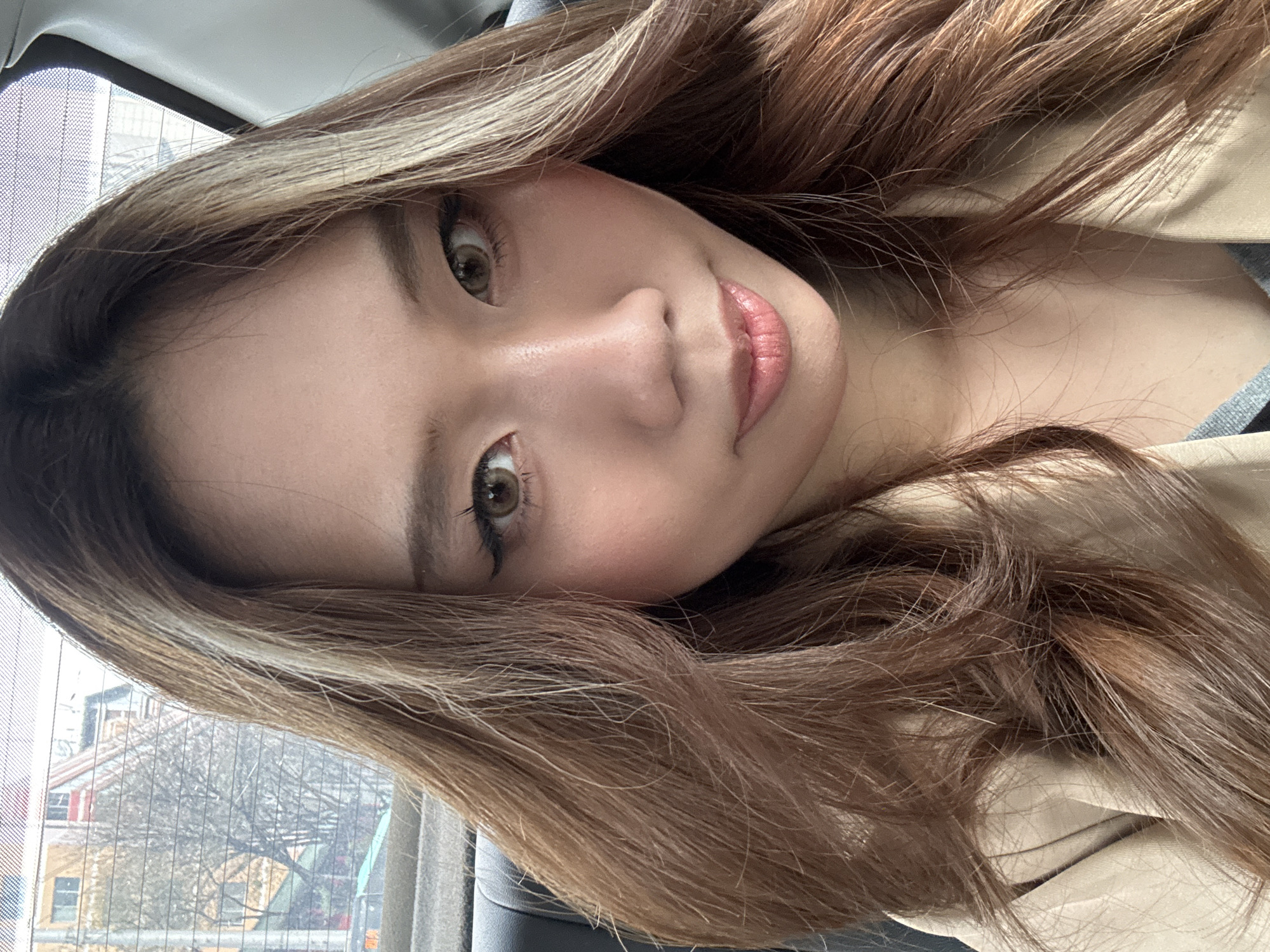 A close-up, slightly tilted portrait of a young woman with light brown hair and blonde highlights. Her skin tone is fair, and she is wearing light pink lip gloss. She has brown eyes and appears to be looking directly at the camera. The background shows a window with a view of buildings and trees. The setting appears to be inside a car or building with a window. There is no information available in the image about the location being in the city of Lanzhou, China.