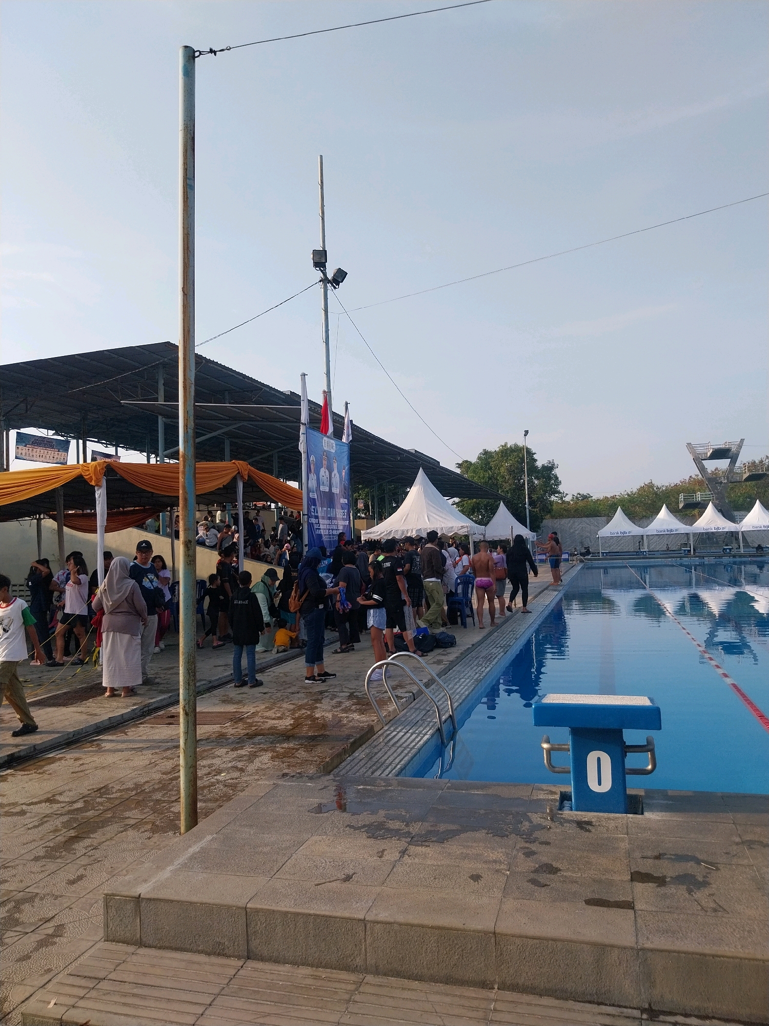 The image depicts an outdoor swimming pool facility, likely in Cirebon, Indonesia, given the context provided. A large, blue swimming pool occupies the right side of the frame, with lane markers visible on the surface. A blue starting block with the number "0" is positioned at the edge of the pool. In the background, several white tents are set up, some bearing the "bank bjb" logo, suggesting a sponsored event. A diving platform is visible further in the distance.
On the left side of the image, a crowd of people are gathered. Some are standing under shaded awnings, while others are milling about on a paved area. Several individuals are dressed in casual attire, and some appear to be in swimwear. There are flags and banners displayed, including one with images of men and text that is partially obscured. A tall, metal pole with wires extends upwards, and another pole with lights is also present. The ground is wet in places, indicating recent activity or cleaning. The sky is clear and bright, suggesting daytime.