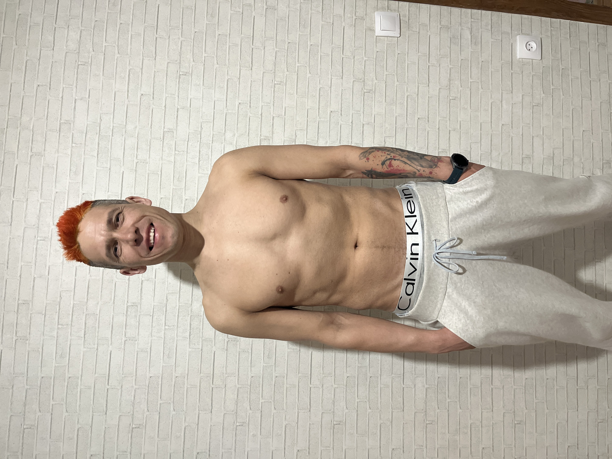 A shirtless man with orange hair, wearing gray Calvin Klein sweatpants and a wristwatch, stands in front of a white brick wall. There are two electrical outlets on the wall, one a standard light switch and the other a European-style power outlet. The location appears to be indoors, potentially a room within a building. There is no information to confirm this is Chaikovsky, Russia.