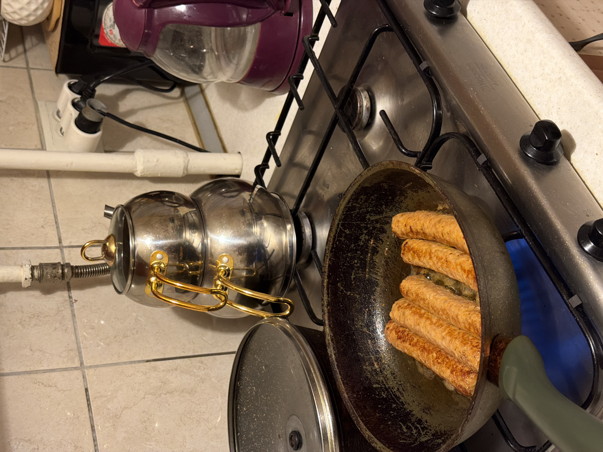 A close-up, eye-level shot captures a stovetop in what appears to be a kitchen. The primary focus is a cast iron skillet on the left, filled with five browned sausages sizzling in oil, with a blue gas flame visible beneath it. To the right of the skillet, a stacked stainless steel teapot with gold handles is positioned on the stove. Further right, a purple electric kettle and a three-outlet electrical adapter plugged into a wall outlet are visible. The background consists of beige tiled walls and a beige countertop. The scene is well-lit, with the overall impression being one of food preparation.