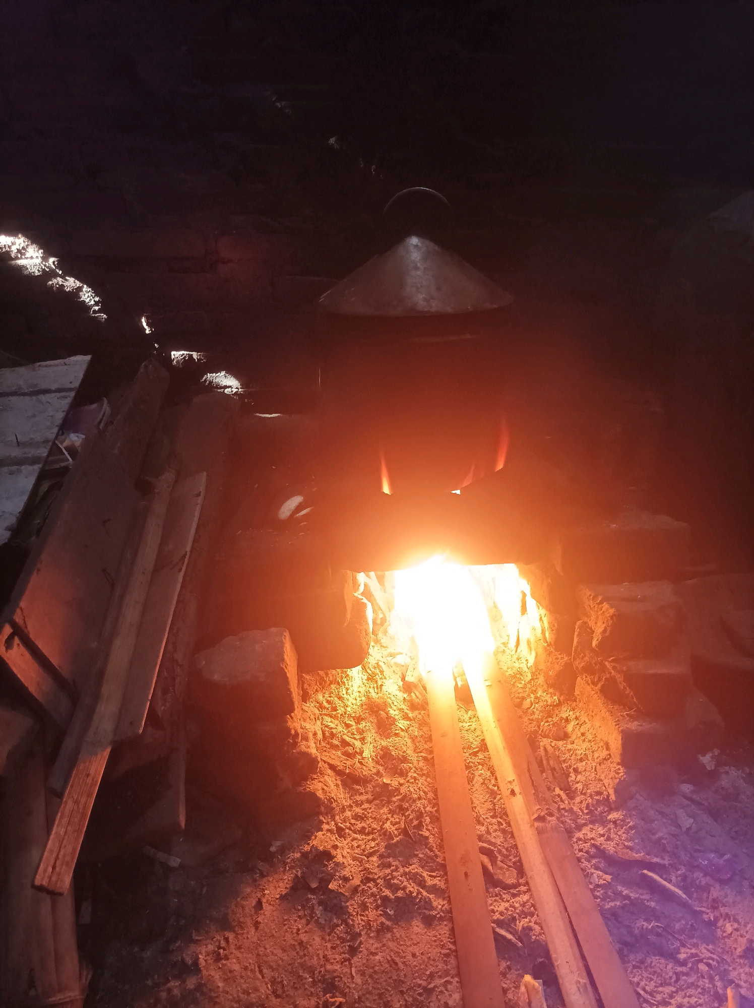 The image depicts an interior scene within a traditional dwelling or workshop in Canga'an, Indonesia. The focal point is a rustic, brick-built furnace or hearth, actively burning with intense orange and white flames. Two long, narrow wooden planks are positioned horizontally within the furnace, appearing to be heated or to be part of a process. Above the opening of the furnace, a conical metal object, possibly a lid or a part of cooking apparatus, is visible. The surroundings are dimly lit, with dark, textured walls or ceilings. To the left of the furnace, a stack of rough-hewn wooden planks is piled up, suggesting construction or fuel materials. The ground appears to be dirt or ash, indicative of the working environment. The overall impression is one of traditional craftsmanship or cooking, utilizing an open flame and natural materials. There are no people visible in the frame.