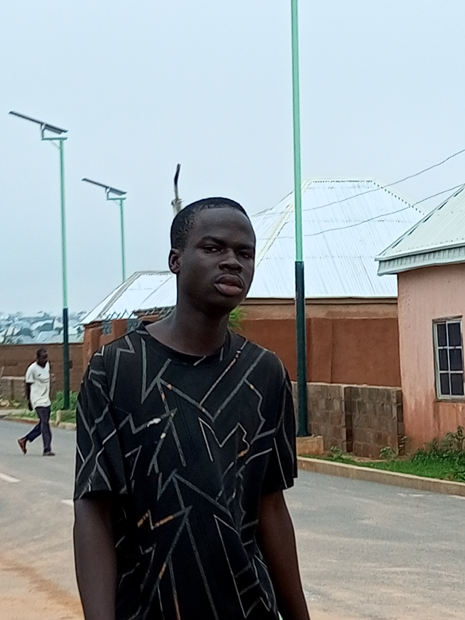 A young Black male stands in the foreground, facing the camera with his body angled slightly to his left. He has dark skin and short, dark hair. He is wearing a black t-shirt with a geometric pattern of thin white and gold lines. His right arm is visible, and his left arm is mostly obscured by his body and shirt.
In the background, a paved road curves to the left. On the right side of the road, there is a pale pink building with a white-capped roof and a single window with a dark frame and multiple panes. Next to this building, there is a wall constructed of brown bricks. Further back, a brown building with a corrugated white roof is visible, and beyond that, a cluster of buildings with similar roofs suggests an urban or semi-urban environment.
On the left side of the road, a street lamp with a solar panel is visible. Another similar street lamp is positioned further down the road. To the left of the road, a person in a white t-shirt and dark pants is walking away from the camera, their back to the viewer. The sky is overcast and light grey. The scene appears to be in an outdoor setting, possibly a residential or developing area within the city of Gombe, Nigeria.