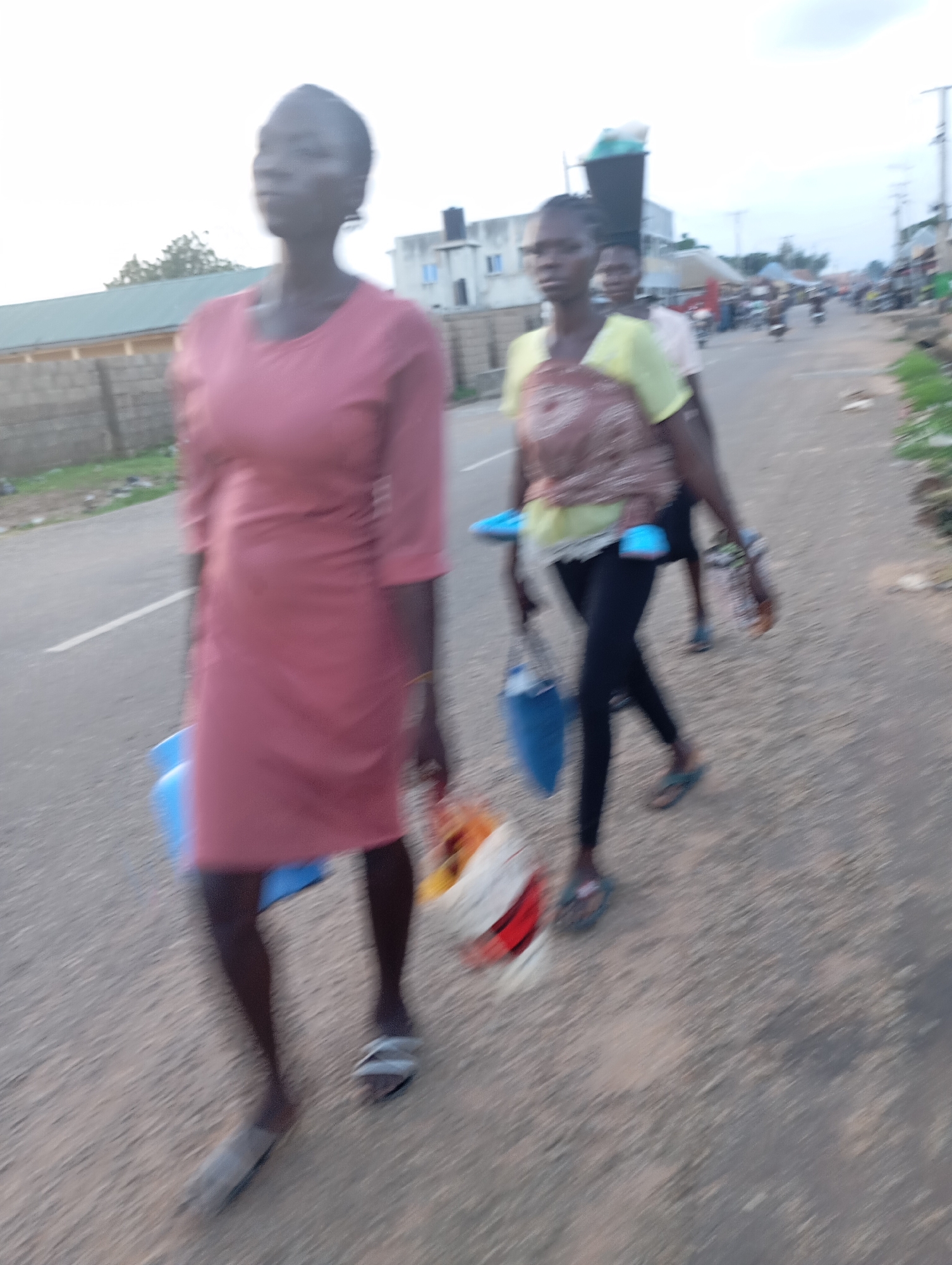 In Shendam, Nigeria, three individuals, appearing to be women, walk along a paved road. The woman in the foreground, on the left, wears a long, rose-colored dress and carries a blue container. To her right and slightly behind, another woman is visible, wearing a light-colored shirt and dark leggings, also carrying a blue bag. A third individual, partially obscured, walks behind them. The road is lined with a low wall and vegetation on the left, and buildings and more vegetation on the right. The sky is overcast. The image is blurred, indicating motion.