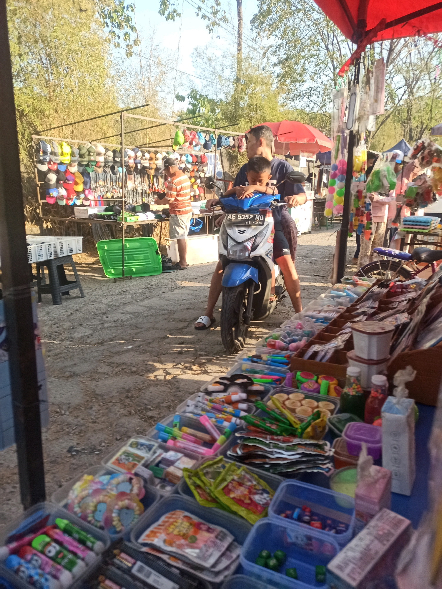 The image depicts an outdoor market scene in Telagamurni, Indonesia. In the foreground, a blue and white motorcycle is parked, with a man in a dark blue shirt and a child in a dark blue t-shirt sitting on it. The man is driving the motorcycle, and the child is seated in front of him. They are both looking to the left. Behind the motorcycle, a stall is set up with a rack of colorful baseball caps. A vendor wearing an orange and white striped shirt and white shorts stands to the right of the caps, facing away from the camera and appearing to be organizing merchandise.
To the right of the motorcycle, a market stall displays an assortment of goods, including what appear to be colorful toys, stationery items, and packaged snacks. The stall is covered by a red canopy. In the background, more market stalls are visible, as well as trees and a bright sky. The ground is covered with dirt and sand. The overall scene suggests a bustling local market on a sunny day.