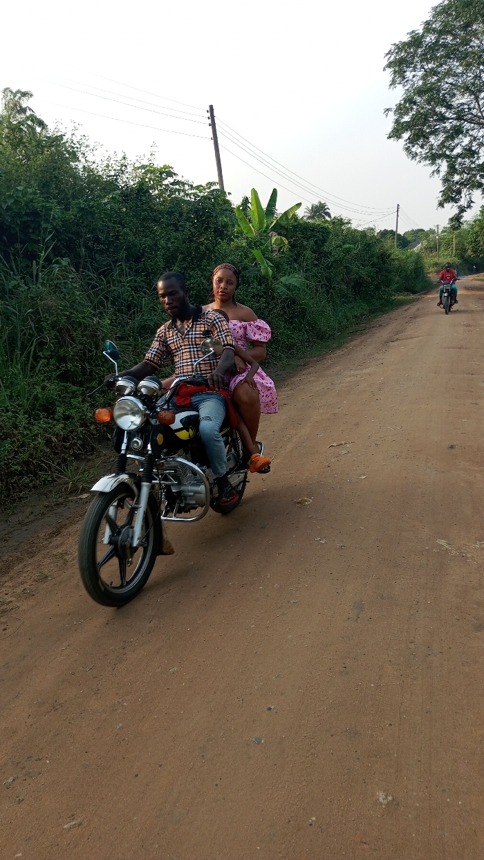A man and woman are riding a black and chrome motorcycle on a dirt road. The man wears a plaid shirt and jeans, while the woman sports a pink floral dress. Green foliage lines the road, and a utility pole stands in the background. Another person is riding a motorcycle in the distance on the same road. The sky is overcast.