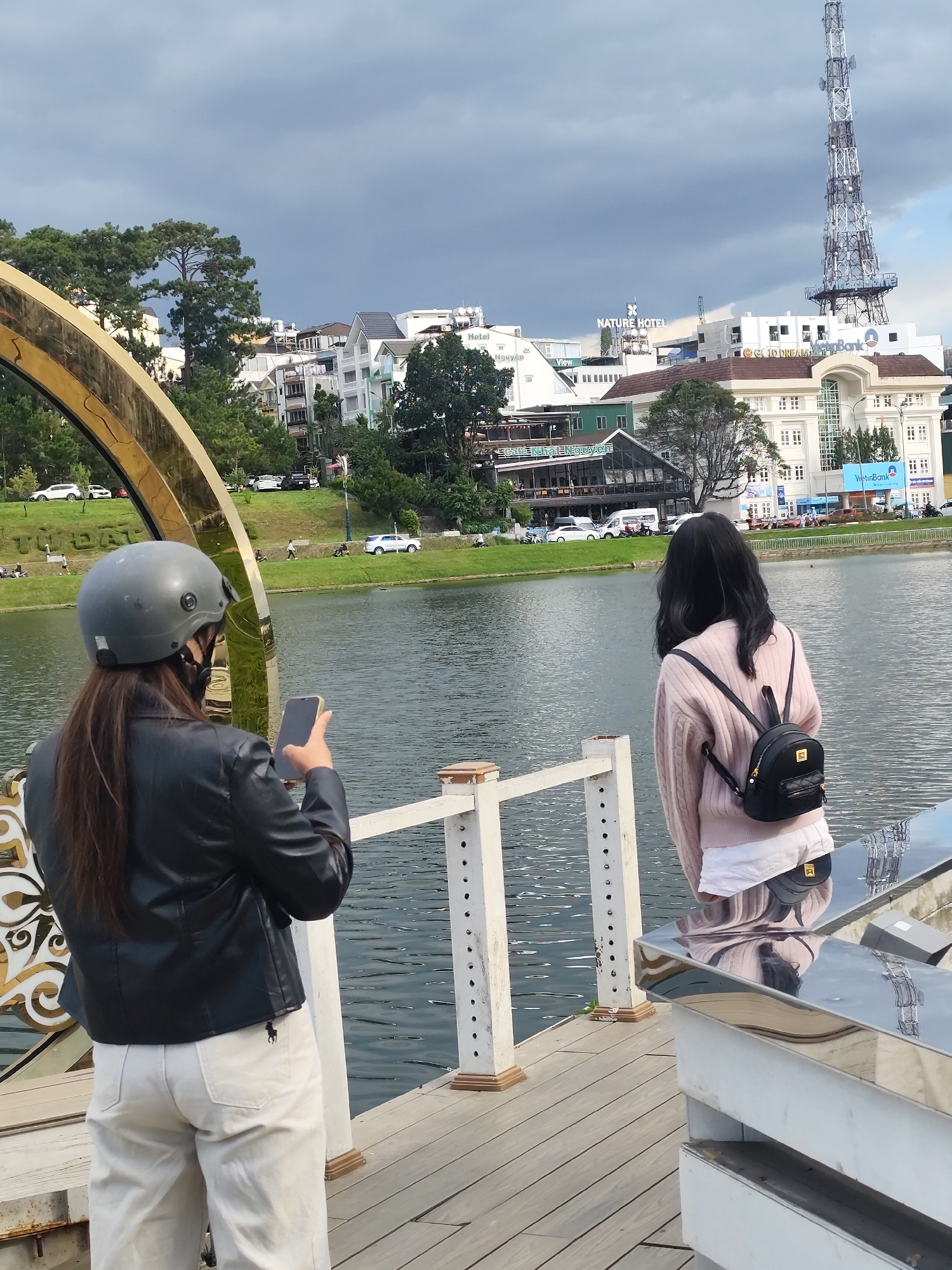 Two individuals are standing on a wooden pier overlooking Xuan Huong Lake in Da Lat, Vietnam. The person in the foreground, wearing a grey helmet, black leather jacket, and light-colored pants, is holding a smartphone and appears to be taking a picture. To the right, another person, viewed from the back, is wearing a light pink sweater over a white top and a small black backpack. They are looking out at the lake. In the background, buildings, trees, and a tall communication tower are visible against a cloudy sky. The lake is calm, reflecting the surrounding scenery. The overall atmosphere suggests a casual outing or sightseeing experience.